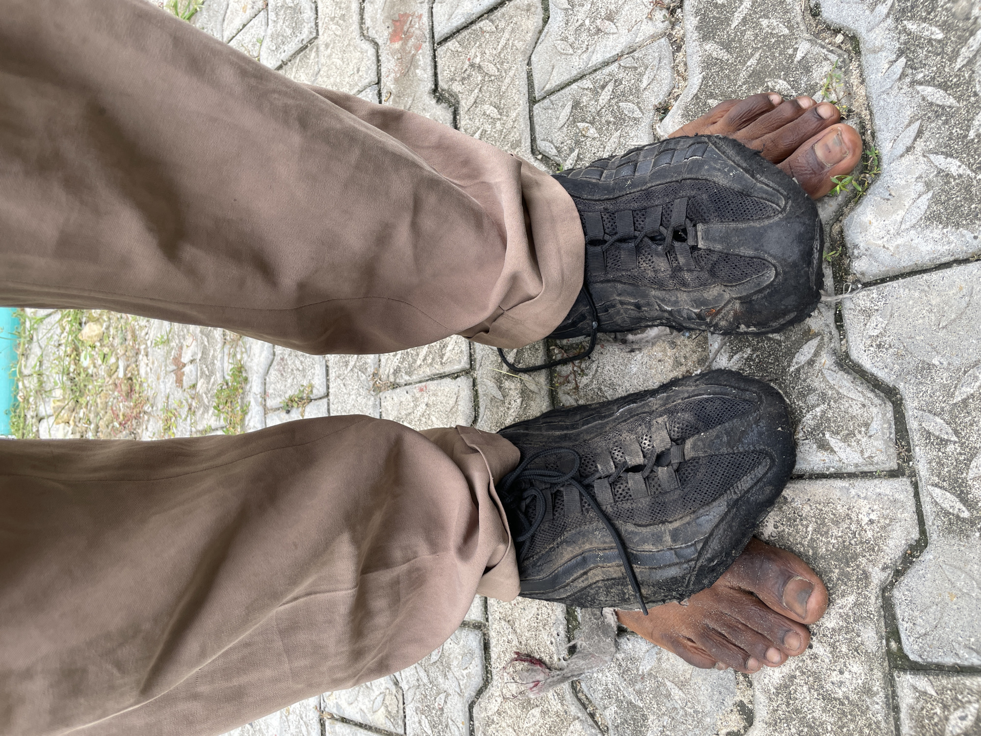 A view from above depicts a person's lower legs and feet standing on a paved surface. The individual is wearing brown trousers with a slightly baggy fit. The person's right foot is partially covered by a worn, dirty black athletic shoe, while the toes are visible. The left foot is similarly adorned with a matching shoe, leaving the toes exposed. The paved ground is composed of gray concrete blocks with a textured, patterned surface. Small sprouts of green vegetation are growing in the crevices between the blocks near the right foot. The location is the city of Ado, Nigeria.