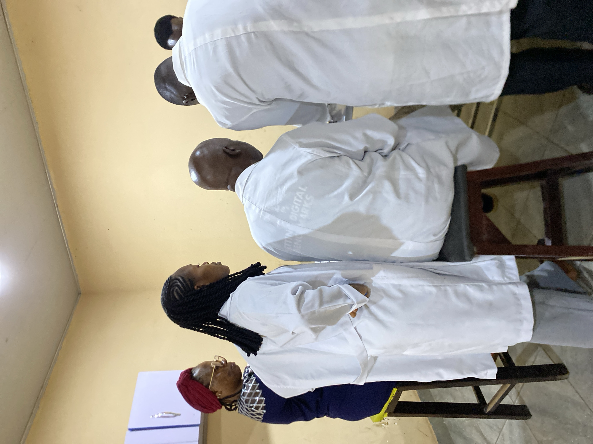 The image shows a group of people in a room, possibly in Bukuru, Nigeria. They are all wearing white lab coats and seated on chairs. Some are wearing casual clothes underneath. The walls are painted yellow and there is a cabinet in the background.