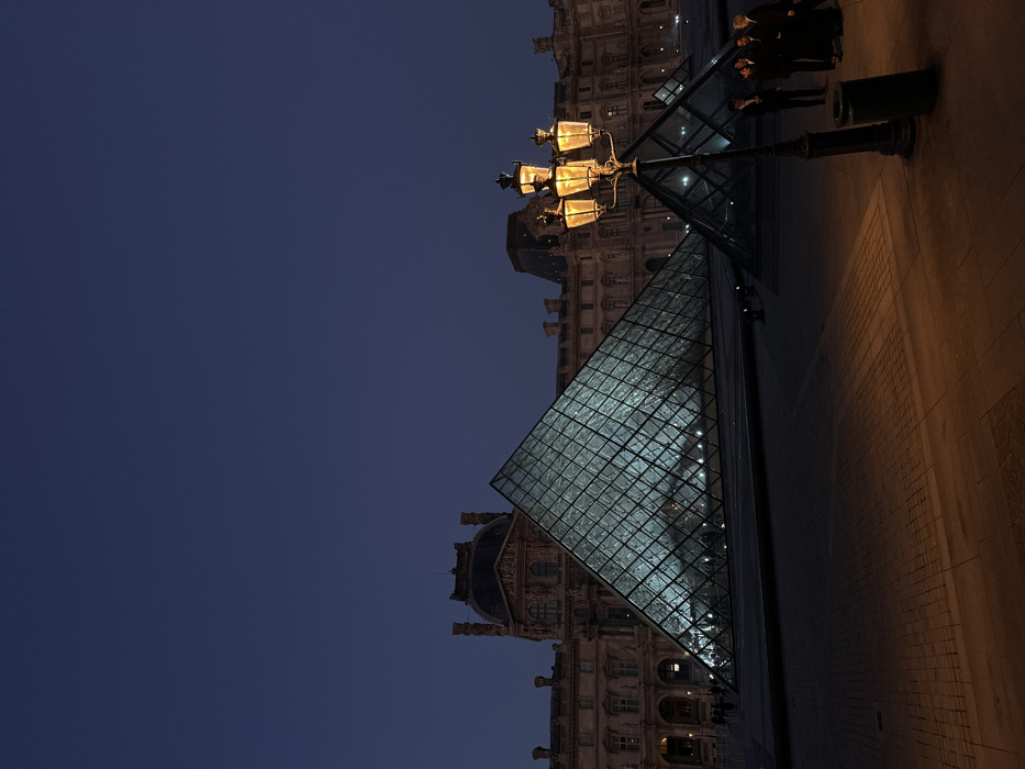 The image depicts the Cour Napoléon of the Louvre Museum in Paris, France, at twilight or night. A large, illuminated glass pyramid, the Louvre Pyramid, occupies the center-left, showcasing its grid-like structure and a teal internal light. Behind it, the classical facade of the Louvre Palace is visible under low illumination against a dark blue sky. To the right of the large pyramid, a smaller glass pyramid is partially visible. In the foreground on the right, a black, ornate lamppost with four lit lanterns emitting an orange glow stands prominently. Beneath the lamppost, a dark, cylindrical trash receptacle is present. A group of approximately five men, dressed in dark clothing, are standing together on the right side of the frame, near the lamppost, appearing to be posing or conversing. The ground consists of patterned paving stones, illuminated by the streetlamp in the immediate foreground. Distant, indistinct figures are faintly visible near the base of the main pyramid on the left.
