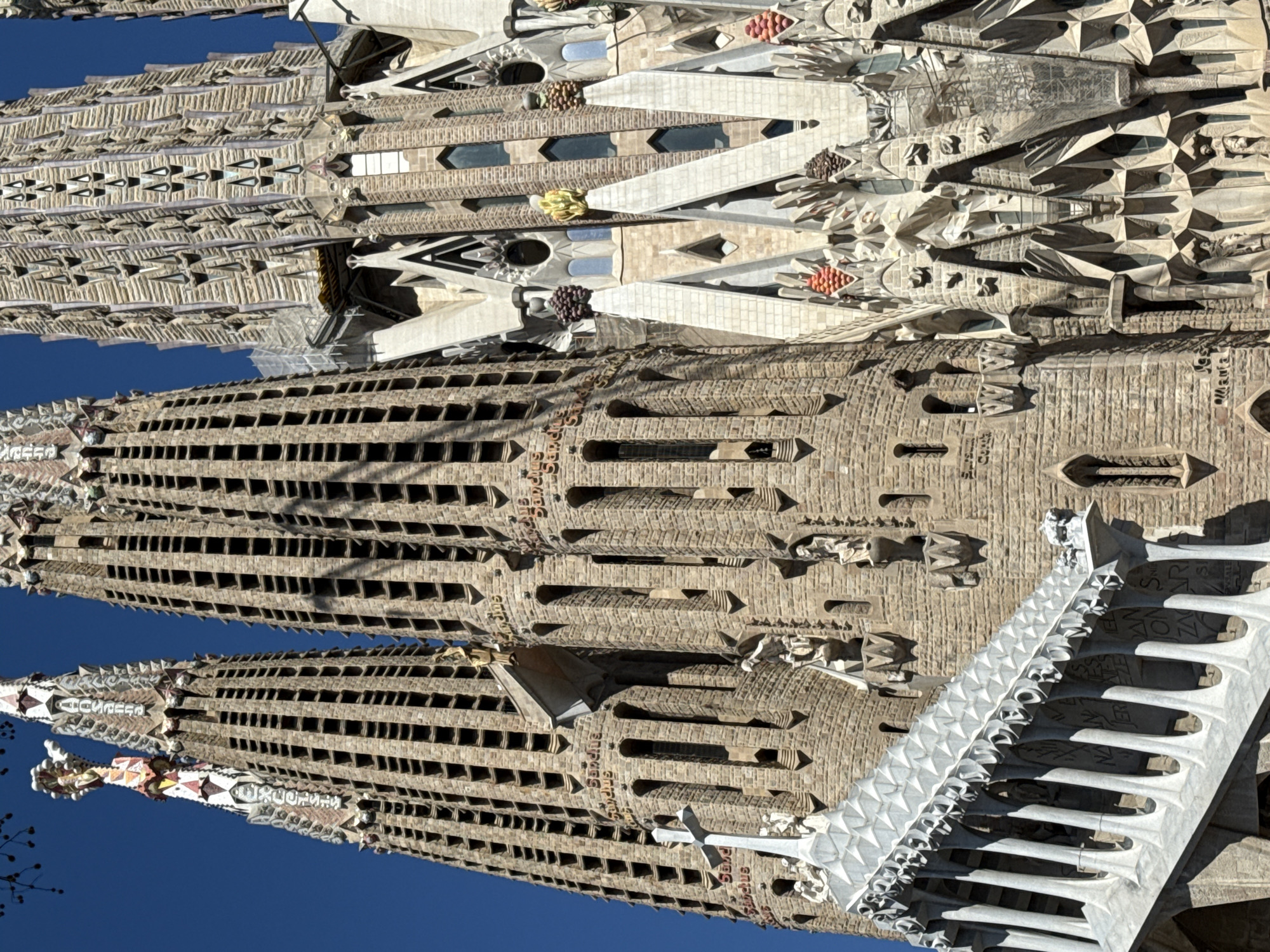 This image showcases a section of the Sagrada Familia in Barcelona, Spain. The view is tilted, focusing on the intricate architectural details of the towers and facade. The towers are characterized by numerous small windows and decorative stone carvings. The upper part of the facade features complex geometric patterns and sculptures. The sky is visible as a clear blue backdrop.