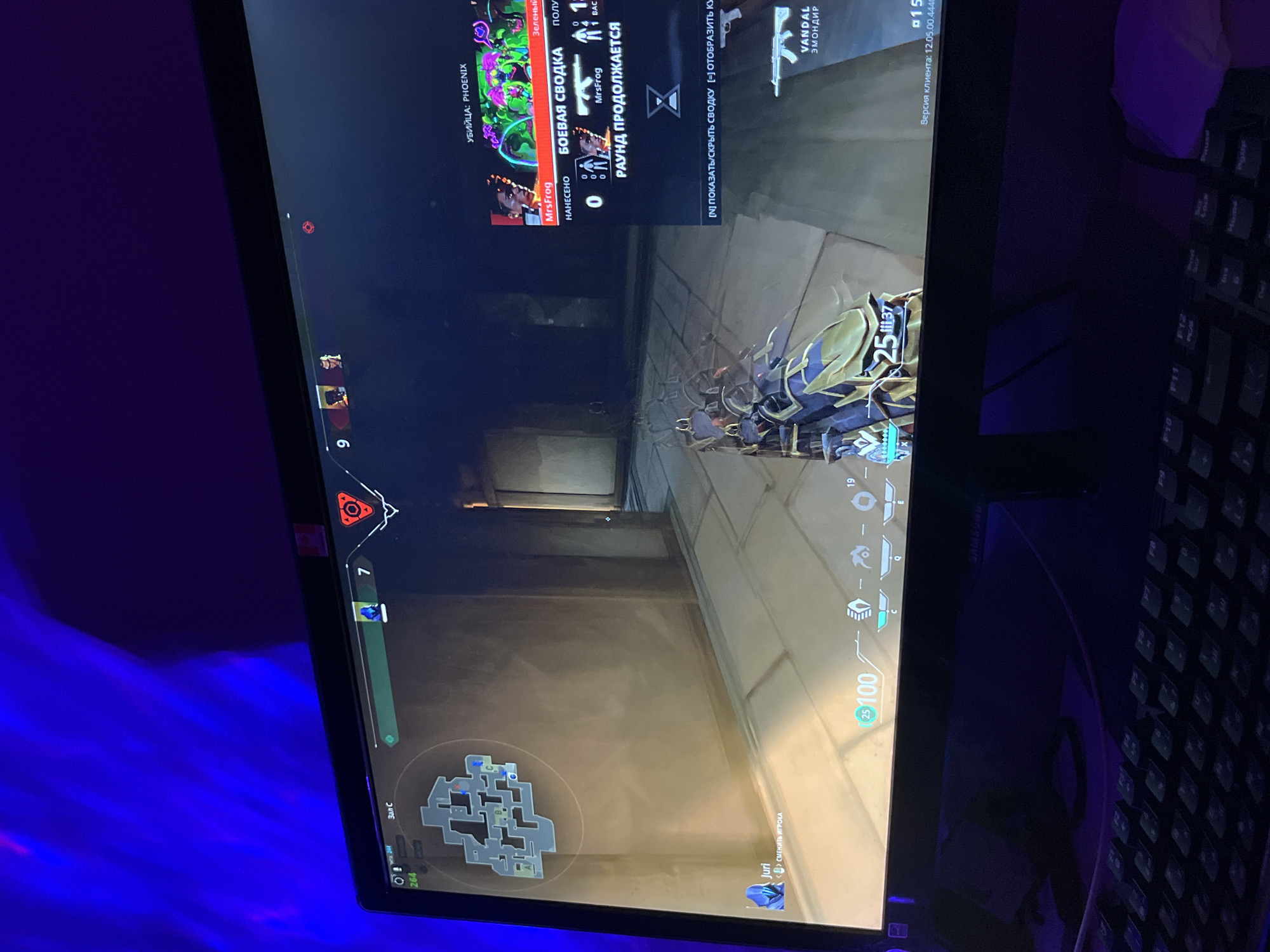 The image shows a computer screen displaying a video game, specifically Valorant. The player is in a round of the game, evidenced by the timer, score, and in-game HUD elements. The player character is visible in the center of the screen, equipped with a weapon (Vandal) as indicated in the top right corner. The mini-map in the bottom left corner shows the game map layout. There is a battle summary on the top left of the screen. The screen is surrounded by a blue ambient light and a keyboard is visible to the right of the screen. There is no information in the image related to the city of Rostov-on-Don, Russia.