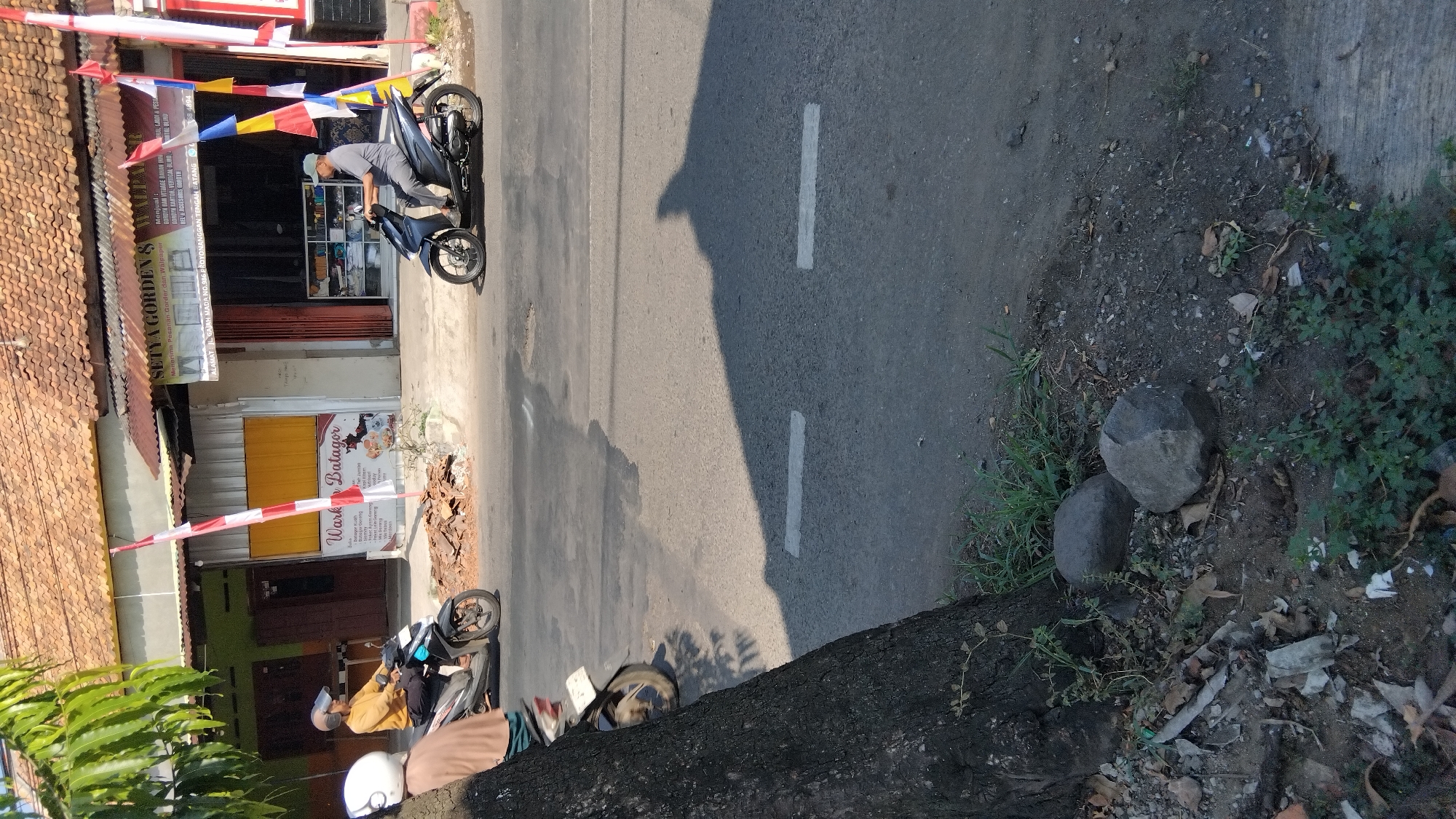 The image shows a street scene in Proyonanggan Tengah, Indonesia, with two motorcycles and their riders. On the left, a person wearing a yellow shirt and helmet is riding a black motorcycle. To the right, a person in a grey shirt and cap is riding a dark blue motorcycle. In the background, there are several shops, one of which has a sign that reads "SETYA GARDEN &" and "Menerima Pesanan Gorden dan Vitrase". Another shop has a sign that says "Wark Batagor". The street is paved, and there are some white lines marking the lanes. The area is lined with trees and vegetation, and there are two rocks visible in the foreground near a large tree trunk. The sun is shining, casting shadows on the road.