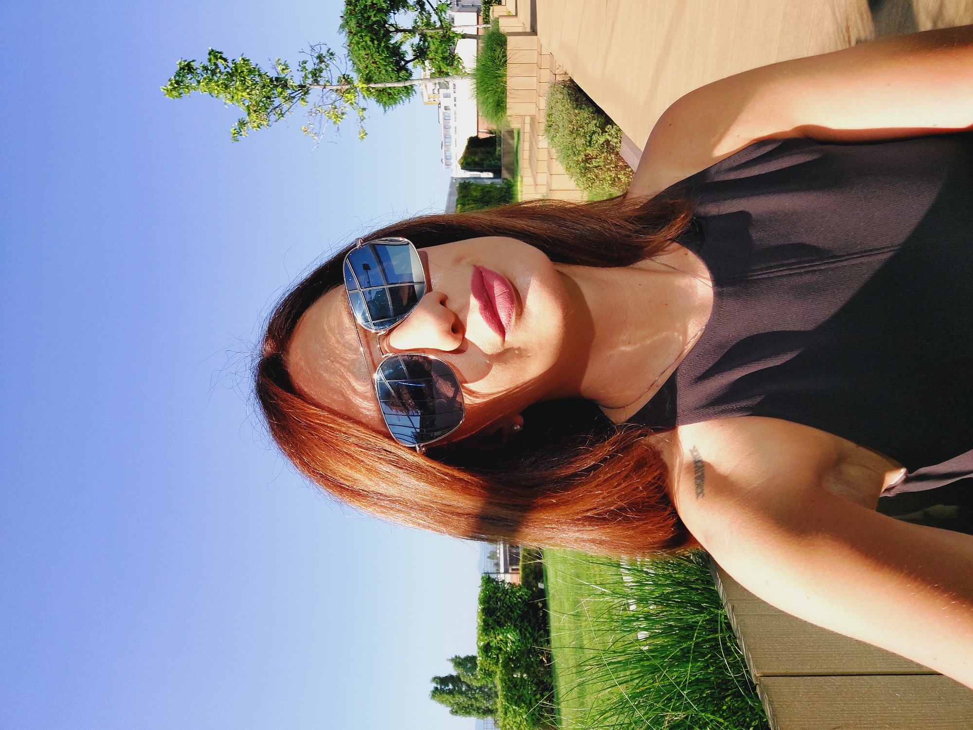 A woman with auburn hair, wearing sunglasses and a black sleeveless top, is posing for a selfie outdoors. She is in the foreground, with a clear blue sky above her. In the background, there is a green lawn, bushes, trees, and some buildings. The scene appears to be a park or a garden in a city. The lighting suggests it is daytime. Based on the provided crops which show a tattoo on the woman's shoulder with what appears to be roman numerals, and the architecture visible in the background, the location is identified as the newly renovated Borisova Gradina Park in Sofia, Bulgaria. Specifically, the area with modern wooden benches and planters is indicative of the "Art Forest" zone within the park.
