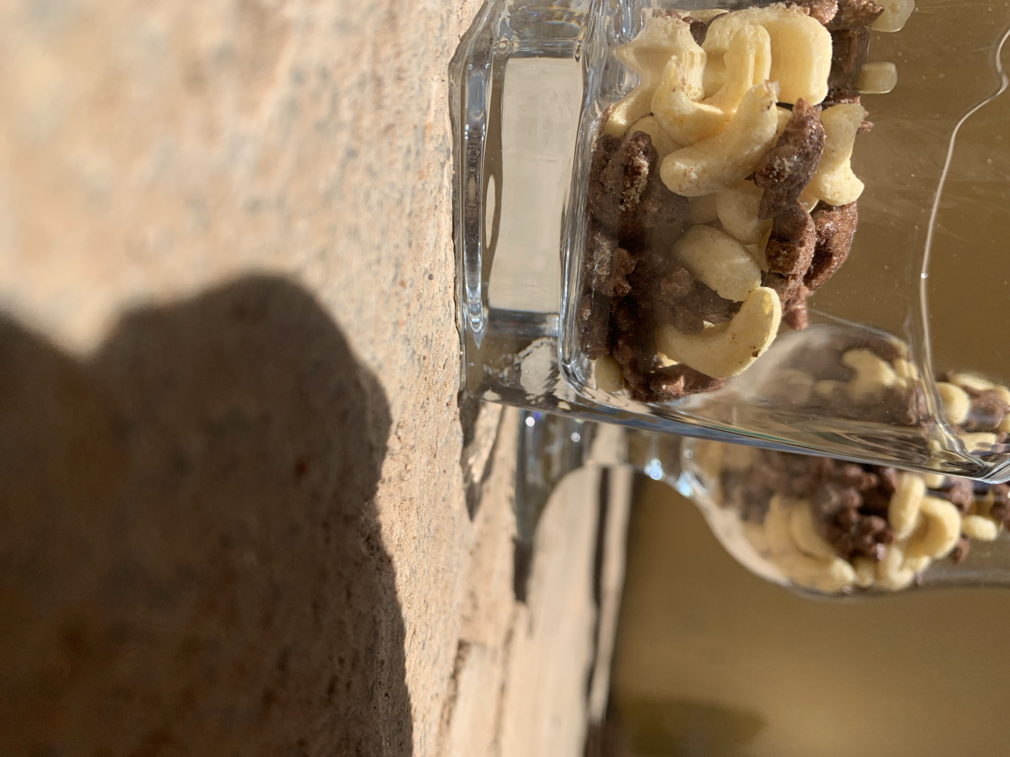 A close-up, ground-level photograph captures two clear glass vessels containing a mixture of light-colored, curved cashew pieces and dark brown, irregularly shaped, granular confectionery or cereal, presented in the city of Rafin Par, Nigeria. The front vessel, a square-based tumbler, is in sharp focus, showcasing the texture and detail of its contents. A second, stemmed glass filled with similar items is visible in soft focus in the background to the right. Both glasses rest on a rough, light brown, outdoor surface, likely stone or paving, bathed in bright natural sunlight which casts a prominent dark shadow across the lower right foreground. The background behind the glasses consists of a blurred, plain tan wall. No individuals are present in the scene.