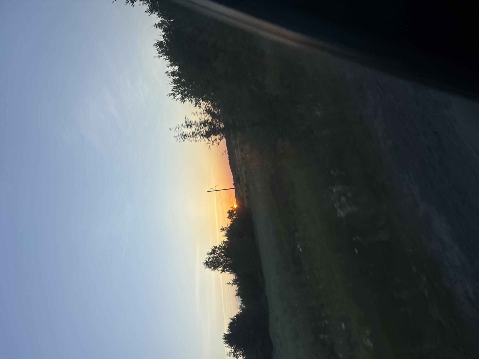 The image depicts a rural scene at sunset, with the sun appearing as a small, bright orange orb low on the horizon. The sky transitions from a clear, pale blue at the top to hues of orange and yellow near the sunset. Silhouetted trees form a dark line across the middle ground, interspersed with a utility pole topped with a single crossbar. To the right of the trees, a grassy field stretches into the distance, appearing darker and less defined due to the fading light. A diagonal dark band, possibly from a window or a car interior, cuts across the upper right portion of the image, suggesting the photo was taken from inside a vehicle. The overall impression is one of a peaceful, natural landscape at the end of the day. The time of day is clearly sunset. The weather appears clear.