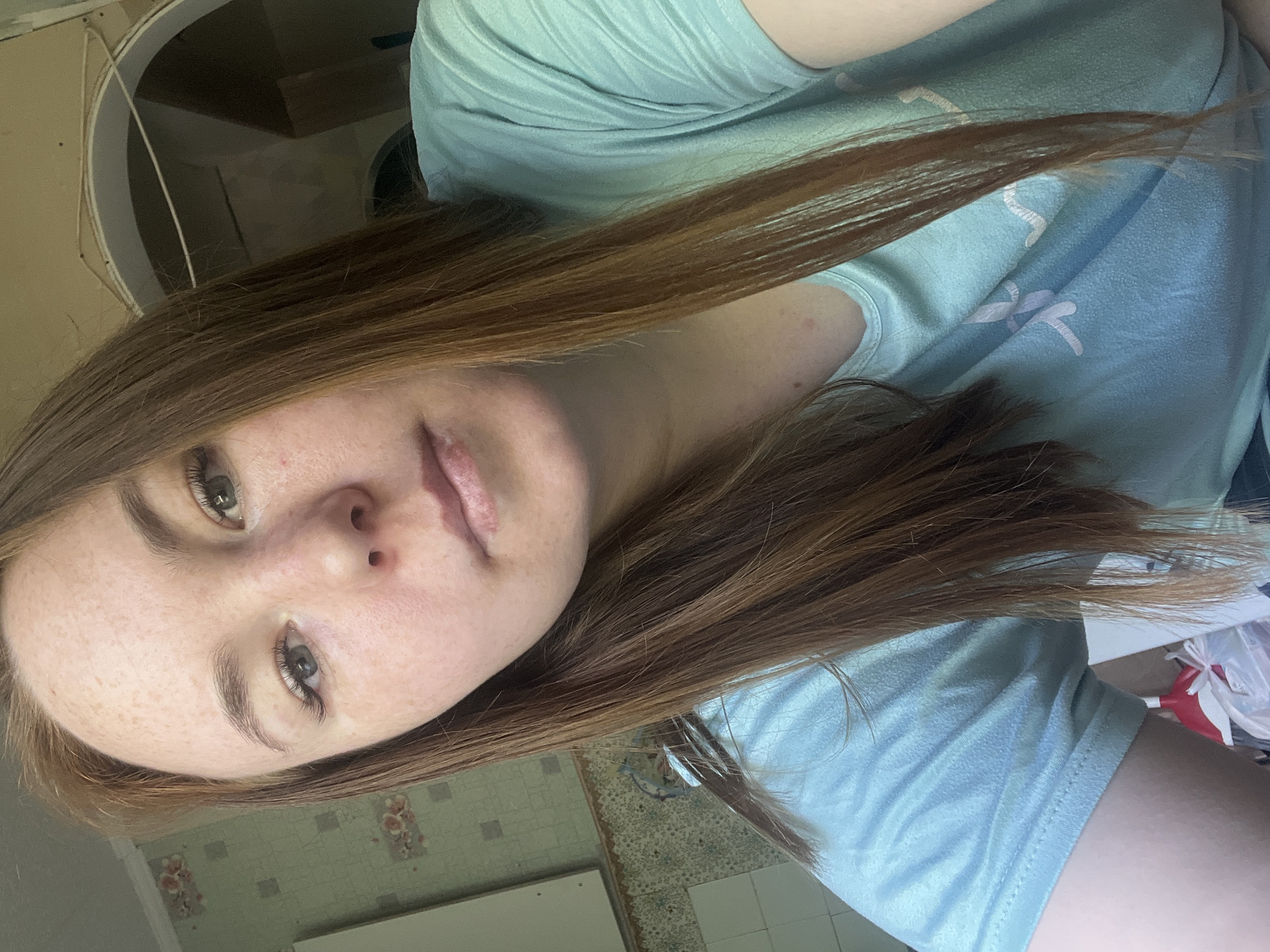 A woman with light brown hair is wearing a light blue t-shirt with a floral design. She has fair skin, freckles, and light-colored eyes. The background appears to be an interior space with tile flooring and potentially a kitchen setting. The location is Chaikovsky, Russia.