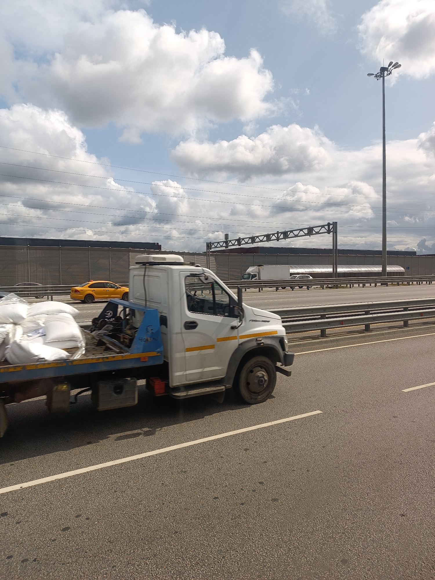 An overhead tow truck is driving on a highway. The truck is white with blue accents and is carrying several large white bags in its bed. In the background, there is a yellow car, a large sound barrier, and a lamppost. The sky is cloudy. The image was taken in Tomilino, Russia.