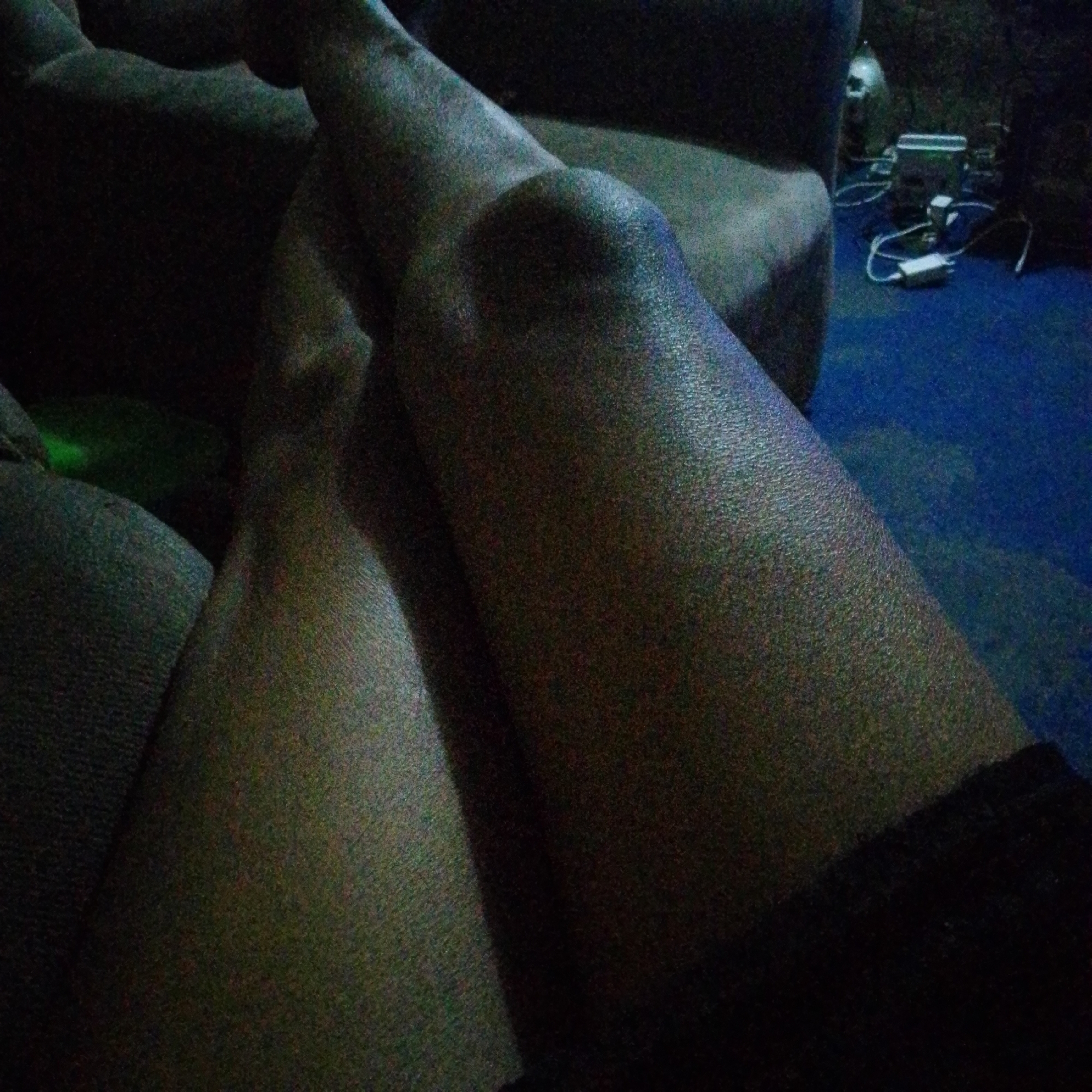 The image shows a close-up of a person's legs resting on a couch. The right leg is in the foreground, with the knee prominently visible. The left leg is partially visible behind it. The person is wearing dark shorts or underwear. In the background, there is a couch, a blue floor, and some electronic devices with wires. The location is Unknown, Unknown.