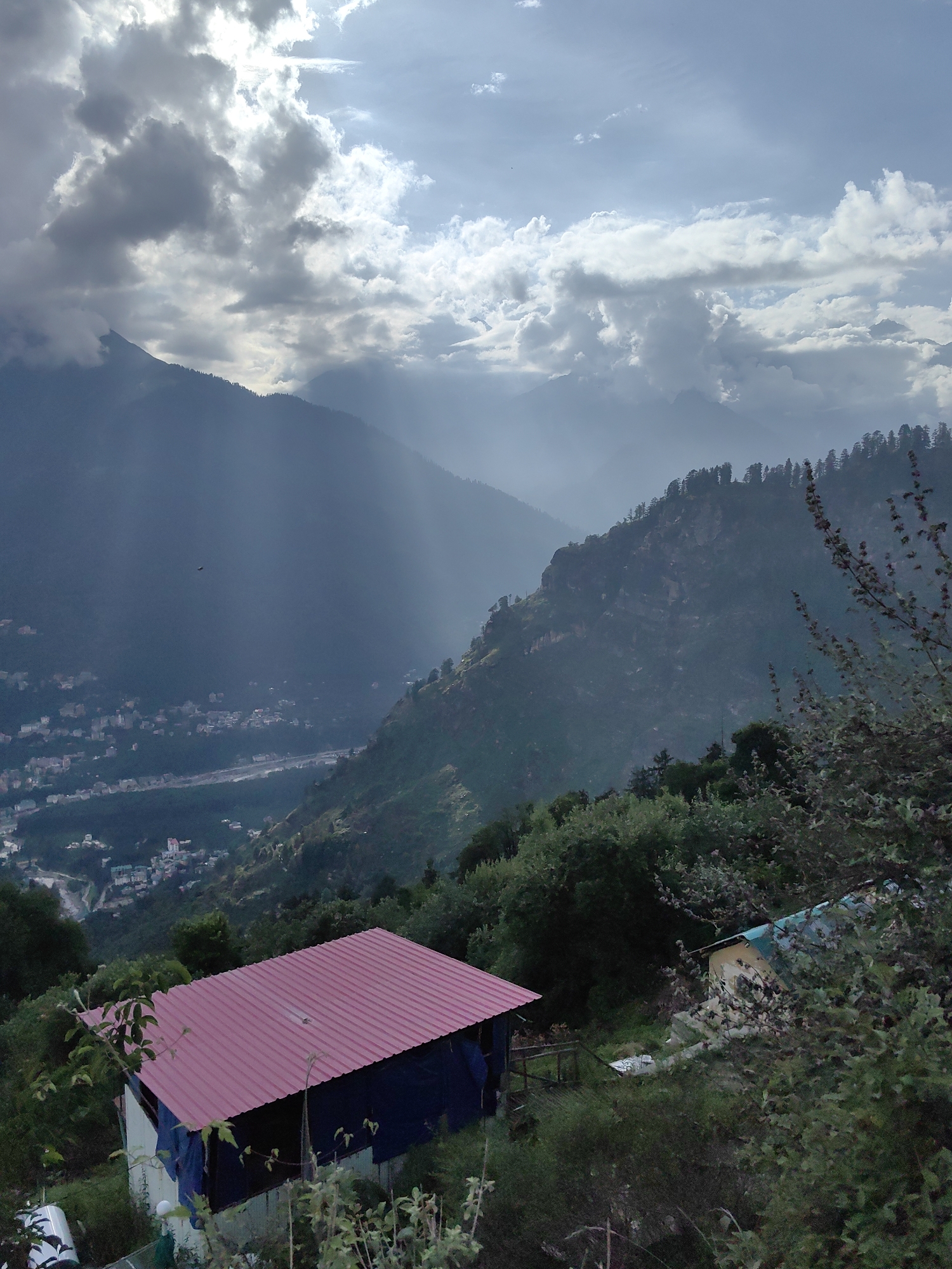The image depicts a scenic mountain landscape with a town nestled in the valley below. In the foreground, there is a dwelling with a red corrugated metal roof and blue tarpaulin walls, surrounded by lush green foliage. To the right, another building with a teal-colored roof is partially visible, also covered in greenery.
The middle ground shows a steep, forested mountainside with a visible path or road winding through it. A small town, identified as being in Aleo, India, is spread across the valley floor, with buildings of various sizes and colors. A river can be seen flowing through the town.
The background is dominated by towering, mist-shrouded mountains. The sky is partially cloudy, with dramatic shafts of sunlight breaking through the clouds, illuminating the valley and mountains. The overall impression is one of natural beauty and a remote, mountainous settlement. There are no people visible in the image.