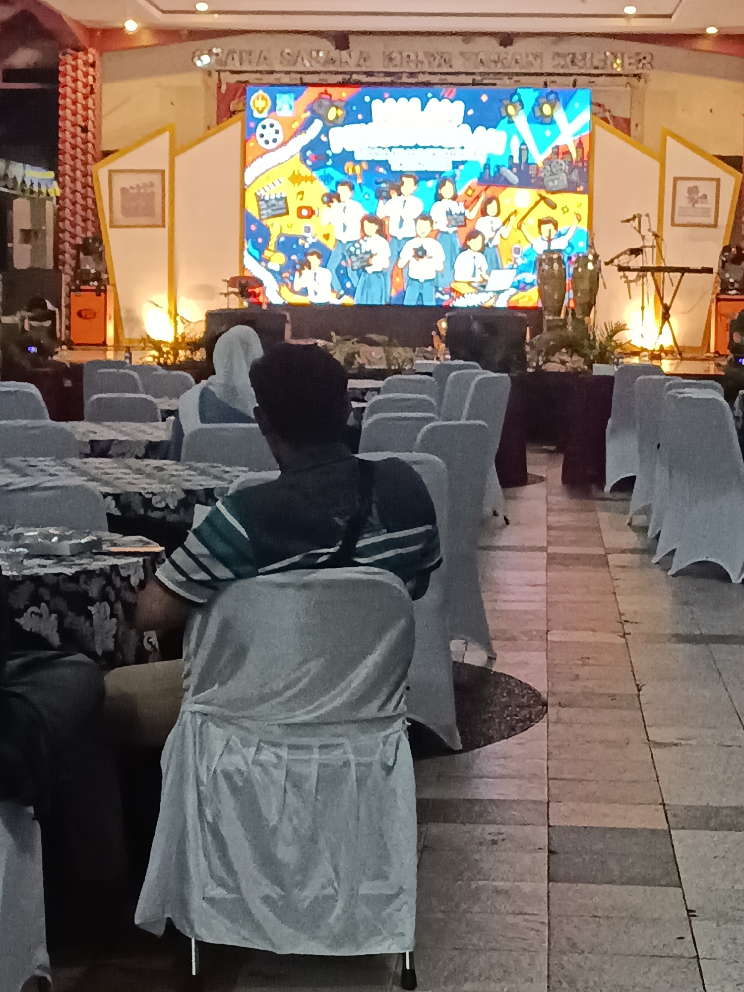 The image shows an indoor event space, likely a hall, in Condongcatur, Indonesia. A large LED screen displaying a colorful, animated graphic is the focal point of the scene. The graphic features cartoonish figures of students, musical instruments, film reels, and cityscapes. In the foreground, several rows of chairs, mostly covered in white fabric, are arranged facing the stage. A few people are seated, with the back of one person in a striped shirt and another wearing a hijab being most prominent. The floor is tiled with a checkered pattern. To the left and right of the stage, there are decorative elements and audio equipment. The overall atmosphere suggests a formal gathering or performance.