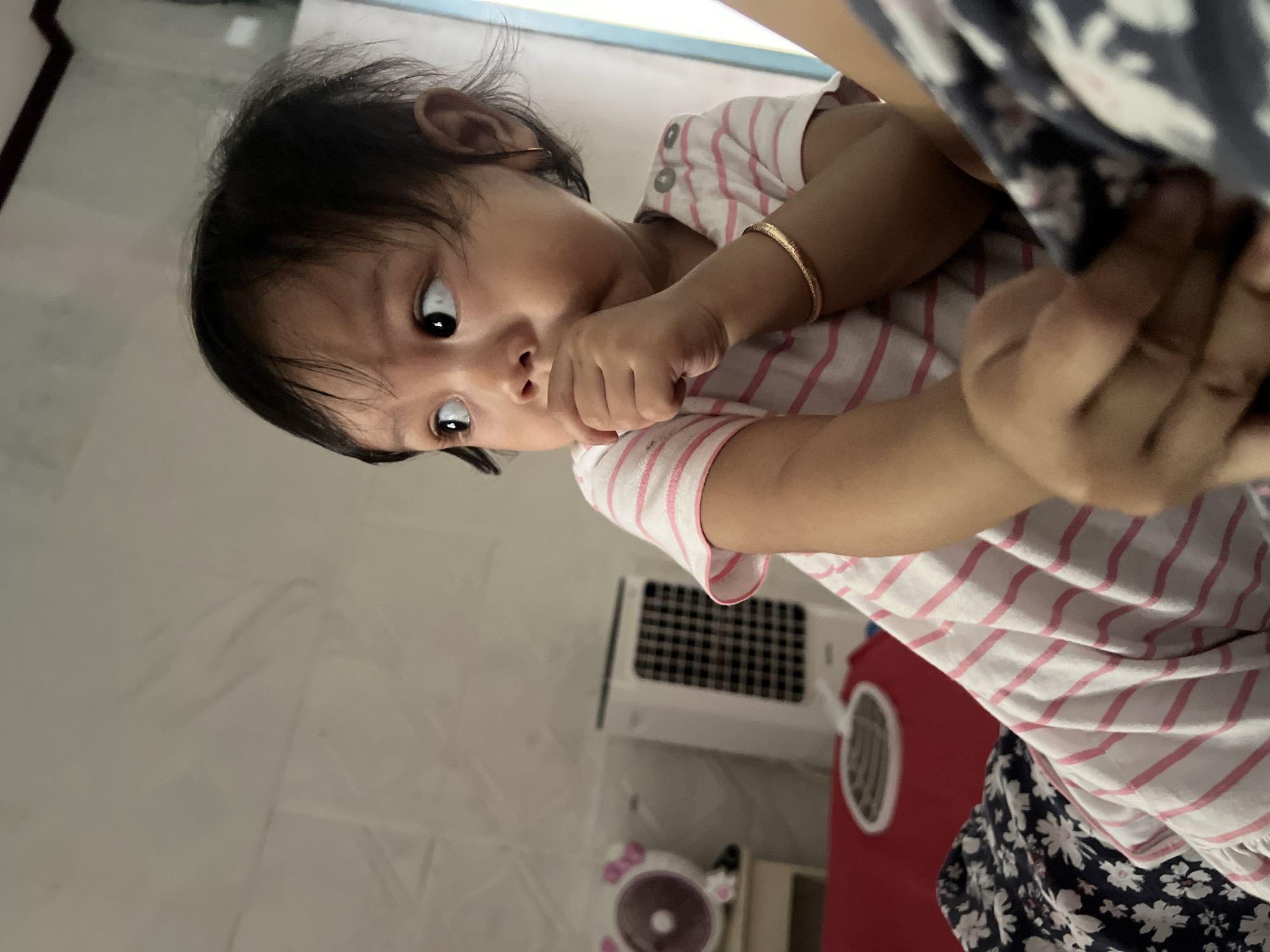 An outdoor shot shows a young child with dark hair and a pink and white striped shirt. The child is looking to the side with their hand near their mouth. The child is wearing a gold bracelet on their right wrist. In the background, there is a white air cooler and a round pink fan. The child is seated on a red surface next to a dark blue blanket with a floral pattern. The image appears to be taken indoors, possibly in a bedroom in Kecamatan Pati, Indonesia.