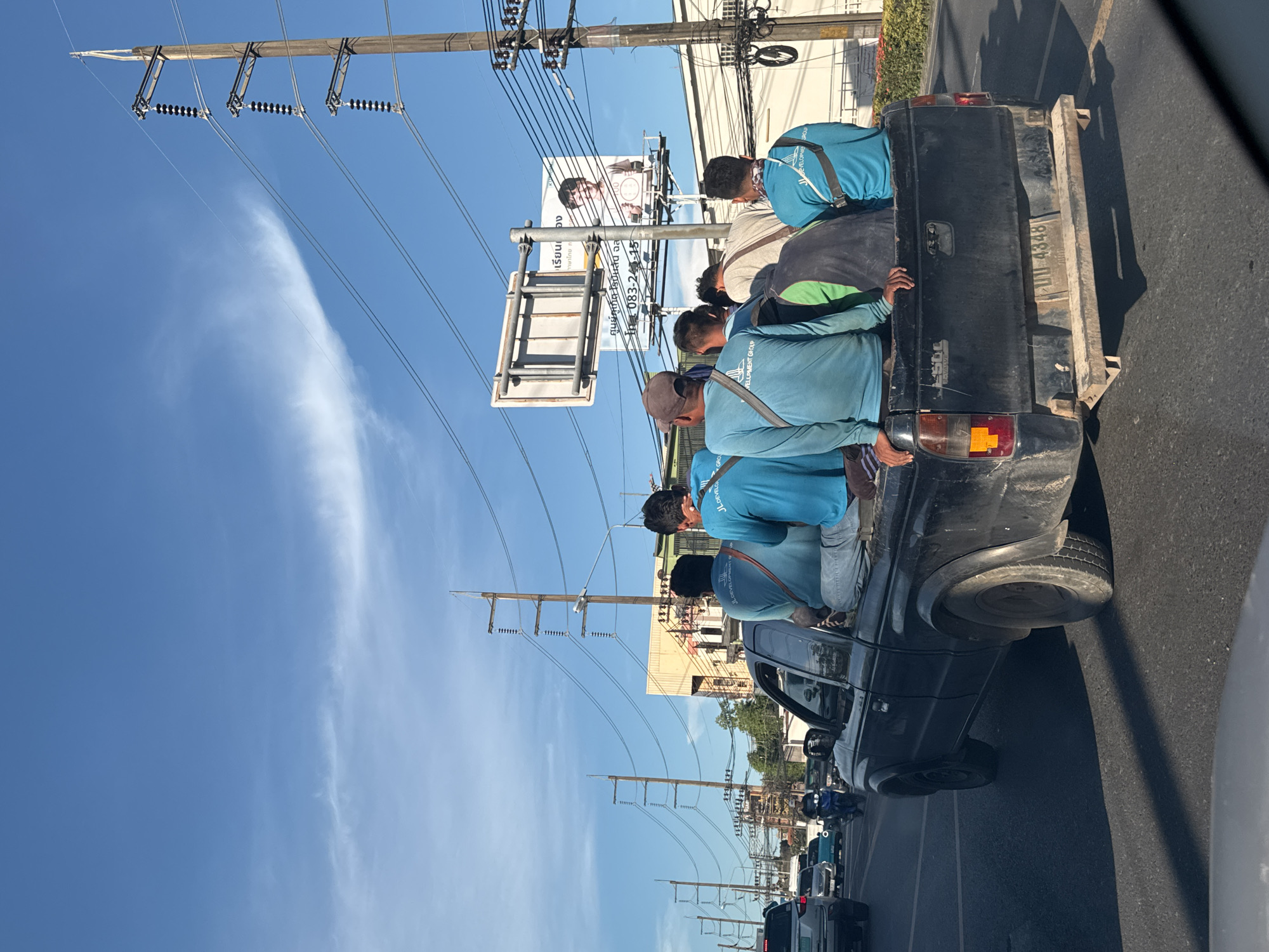 A black pickup truck is driving down a road, carrying several men in the bed of the truck. The men are all wearing blue shirts and appear to be laborers. The truck is in motion, and there are other vehicles on the road as well, including cars and motorcycles. There are power lines and utility poles along the side of the road, and a billboard is visible in the background. The sky is blue with some clouds. The scene appears to be in an urban or suburban area. The license plate on the truck reads "UU 4348".
