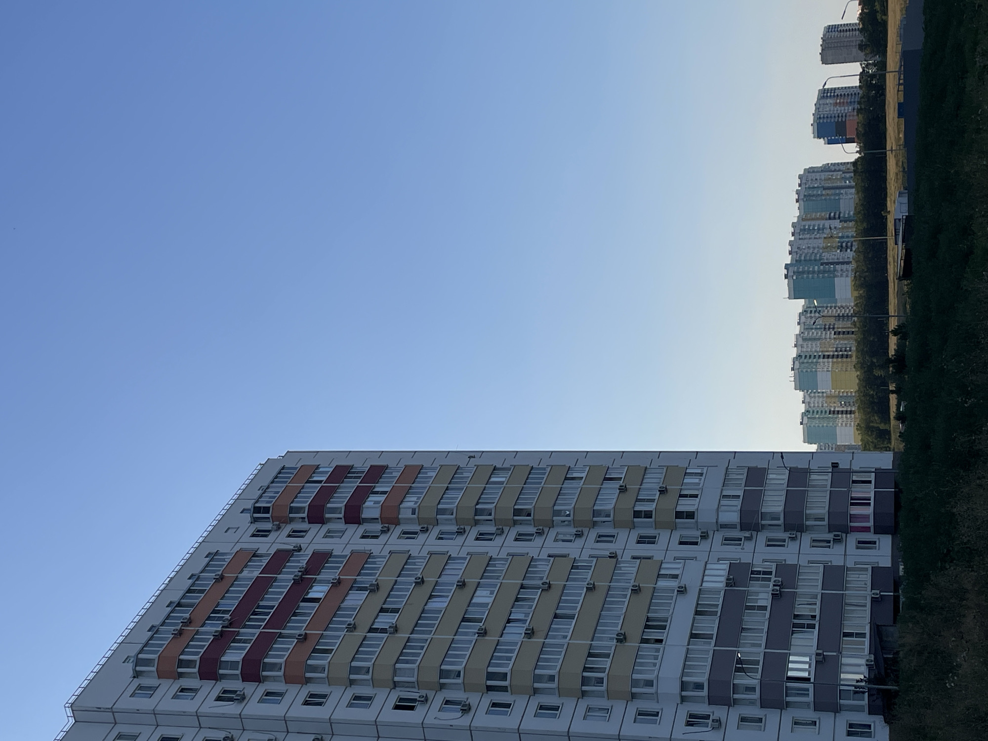 The image depicts a clear blue sky above a residential area in Rostov-on-Don, Russia. In the foreground, a tall, multi-story apartment building with white and colored sections is visible. The building has numerous windows and balconies, many of which are equipped with air conditioning units. To the right and in the background, several other apartment buildings of varying heights and colors can be seen, some with prominent blue, yellow, and red facades. A line of trees separates these buildings from a grassy foreground. The overall scene suggests a modern, developing urban landscape under a bright, cloudless sky. There are no people or animals visible in the image.