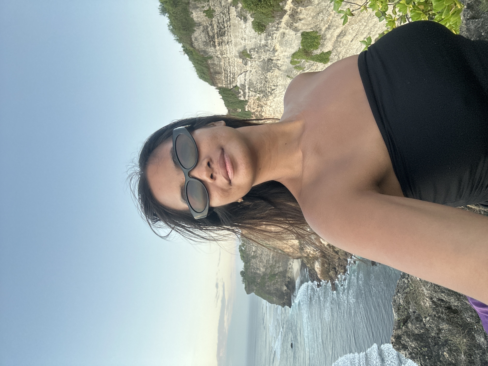 A medium shot captures a woman wearing black sunglasses and a black strapless top, posing for a selfie on a cliff overlooking the ocean. Her dark hair is windswept. Behind her, a rocky cliff face is covered in greenery, and the ocean stretches out to the horizon with waves crashing against the shore. The sky above is a clear, pale blue. The scene appears to be at Uluwatu Temple in Kuta Selatan, Indonesia, known for its dramatic cliffside location.
