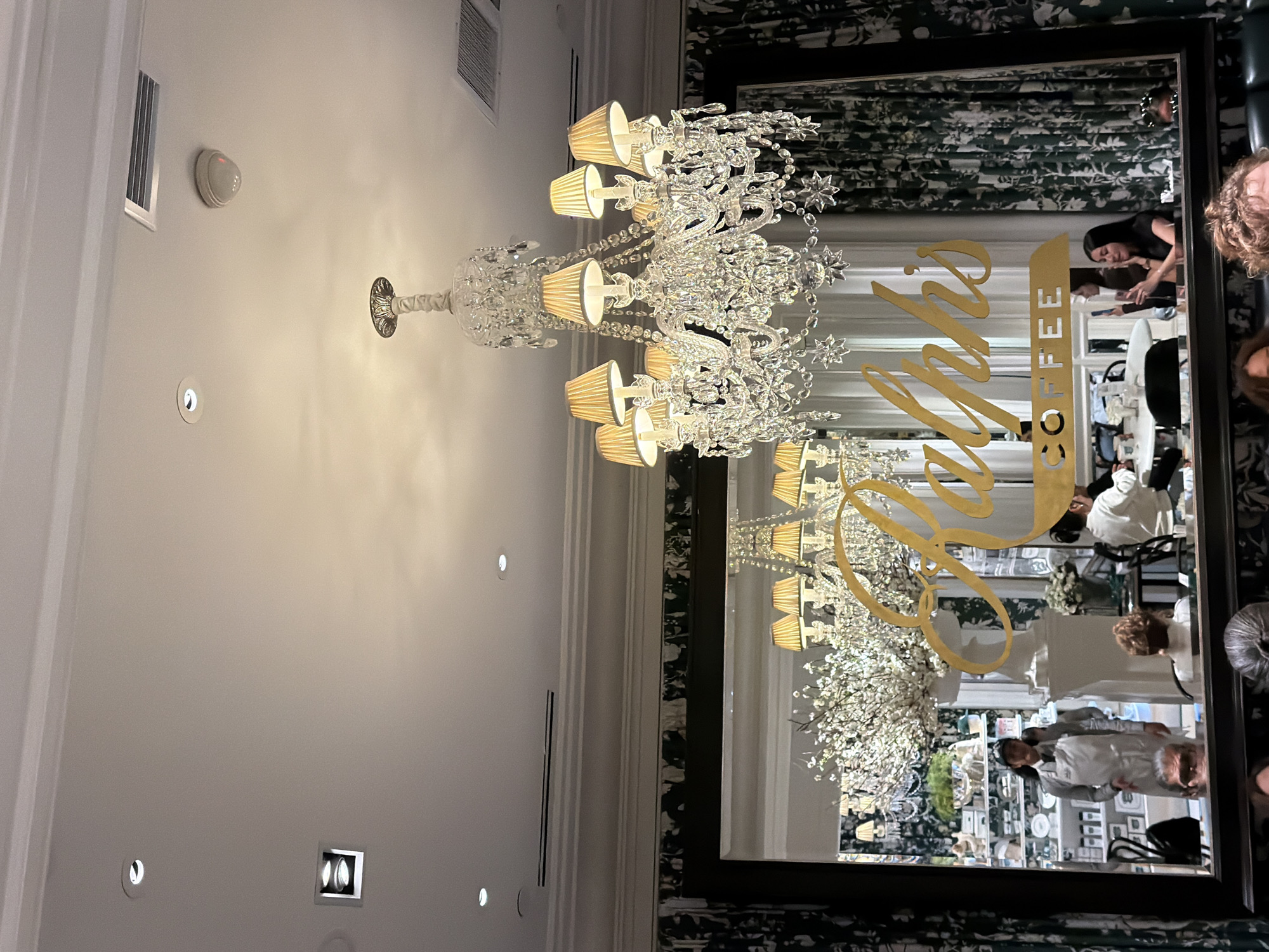 The image displays the interior of Ralph's Coffee, located at 888 Madison Avenue, New York, United States of America. A large, multi-tiered crystal chandelier with ten arms, each bearing a light with a beige lampshade, hangs prominently from the white ceiling. The ceiling is equipped with various recessed lighting fixtures (both circular and square), rectangular air vents, and a white smoke detector.
A large, dark-framed rectangular mirror occupies the mid-ground, reflecting elements of the establishment. The golden, stylized cursive text "Ralph's" is prominently displayed on the mirror's surface, positioned above a golden rectangular sign with white text reading "COFFEE." Within the mirror's reflection, a male staff member wearing a dark cap, a light-colored apron, and a dark tie stands near a large white floral arrangement on a pedestal, which obscures part of a white shelving unit displaying ceramic items. Several patrons are visible seated at tables in the reflection, including a female with long dark hair holding a smartphone. Dark green curtains with a white floral pattern flank the mirror on both sides, and also appear in the mirror's reflection. The tops of two patrons' heads are visible in the immediate foreground at the bottom of the image.