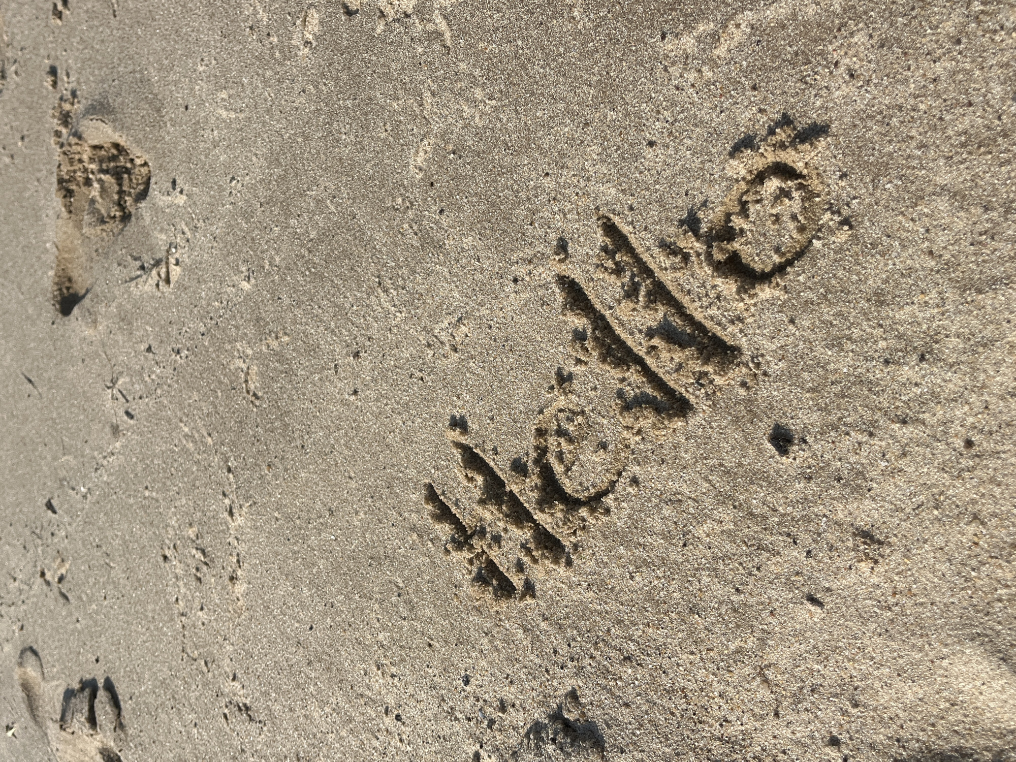 The image shows the word "hello" written in the sand on a beach. The sand is light brown and appears to be damp. There are some footprints in the sand in the upper part of the image. The lighting suggests it is daytime. The location is not specified in Nizhny Novgorod, Russia.