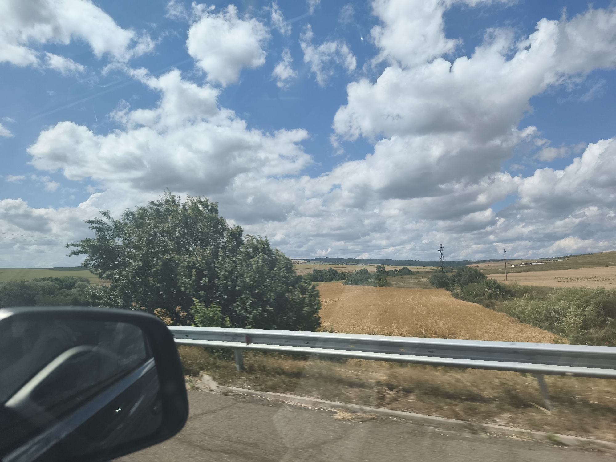 The image depicts a rural landscape viewed from a vehicle, possibly a car, as evidenced by the reflection in the side mirror on the left. The foreground shows a road and a metallic guardrail. Beyond the guardrail, a field of golden crops, likely wheat or barley, stretches into the distance. A large, dense tree with green foliage stands prominently to the left of the crop field. The background features rolling hills with scattered vegetation and a few distant structures, including what appears to be a power pylon. The sky is a vibrant blue, populated with fluffy white cumulus clouds. There are faint contrails from an aircraft visible in the sky. The environment suggests a sunny day in a temperate region, with the crops appearing ripe for harvest. The scene is consistent with an agricultural area. The user has not provided information about the specific location being the city of Troyanovo, Bulgaria, therefore, it cannot be included in the description.