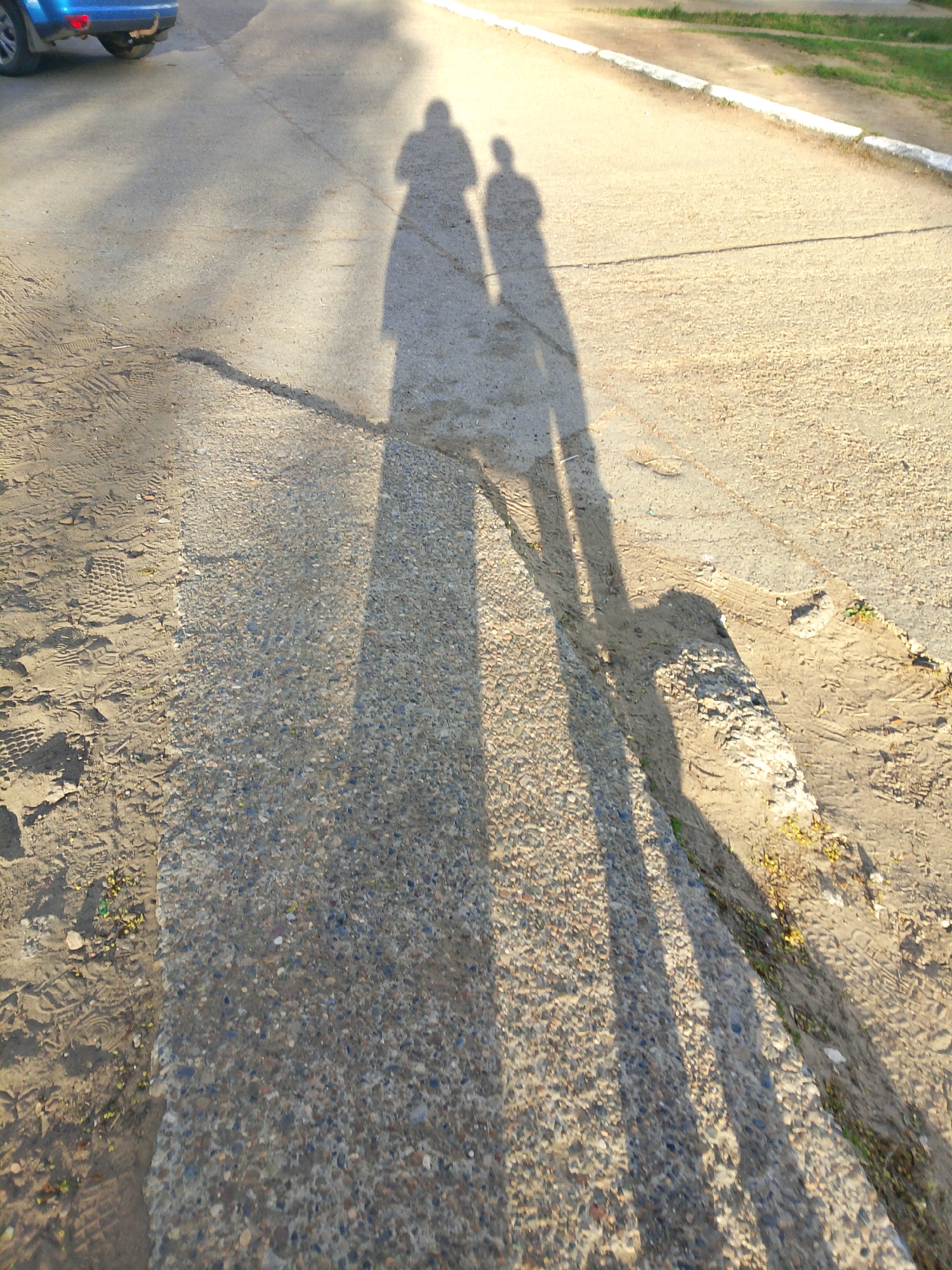 The image shows a street scene on what appears to be a paved road in Noviy, Russia. The surface of the road is a mix of concrete and gravel, with tire tracks and footprints visible in the sand along the side. The shadows of two people are cast onto the road surface, suggesting a sunny day. The road is bordered by grass and a curb. A portion of a blue vehicle is visible in the upper left corner of the frame.