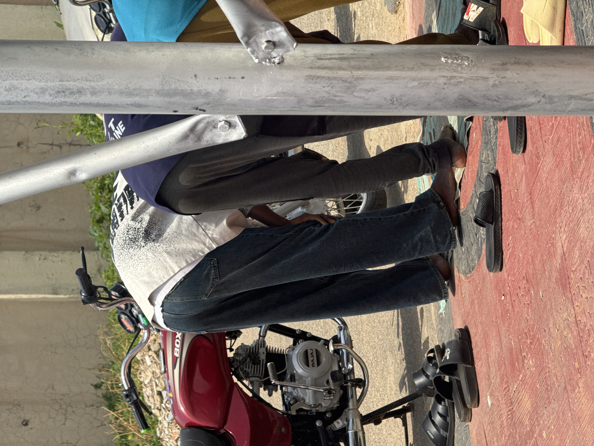 The image captures a close-up view of several people, their lower bodies and feet visible, as they sit or stand near a red motorcycle. The subjects are dressed casually, with one person wearing a white t-shirt with black graphic print and dark blue jeans, another in dark grey trousers, and a third partially visible wearing a blue top. Various footwear, including black slides and sandals, are scattered on the ground. The scene appears to be outdoors, possibly a street or roadside, with a red and black patterned surface visible in the foreground and a concrete wall in the background. The motorcycle is a prominent object, with its handlebars and engine clearly visible. The overall impression is one of a casual gathering or moment of rest. The lighting suggests it is daytime, with strong sunlight creating shadows. There are no explicit clues about the weather, but the outdoor setting implies fair conditions. A partial view of the word "BOX" is visible on the motorcycle. The context of Abuja, Nigeria is provided, which could hint at the cultural environment.