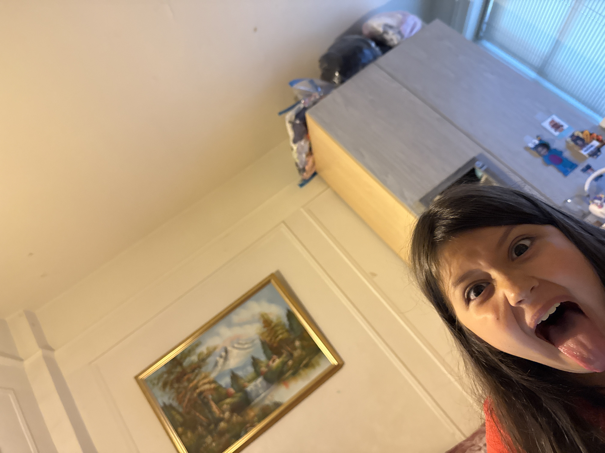 A young girl with long, dark hair and wide, dark eyes is making a funny face, sticking her tongue out at the camera. She is wearing a red top. The background shows a cream-colored wall with molding, a framed painting with a landscape scene, and a grey cabinet with some objects on top, including what appear to be bags of items. There are also blinds on a window in the background. The scene appears to be indoors, possibly in a home. The location is not specified in the image.