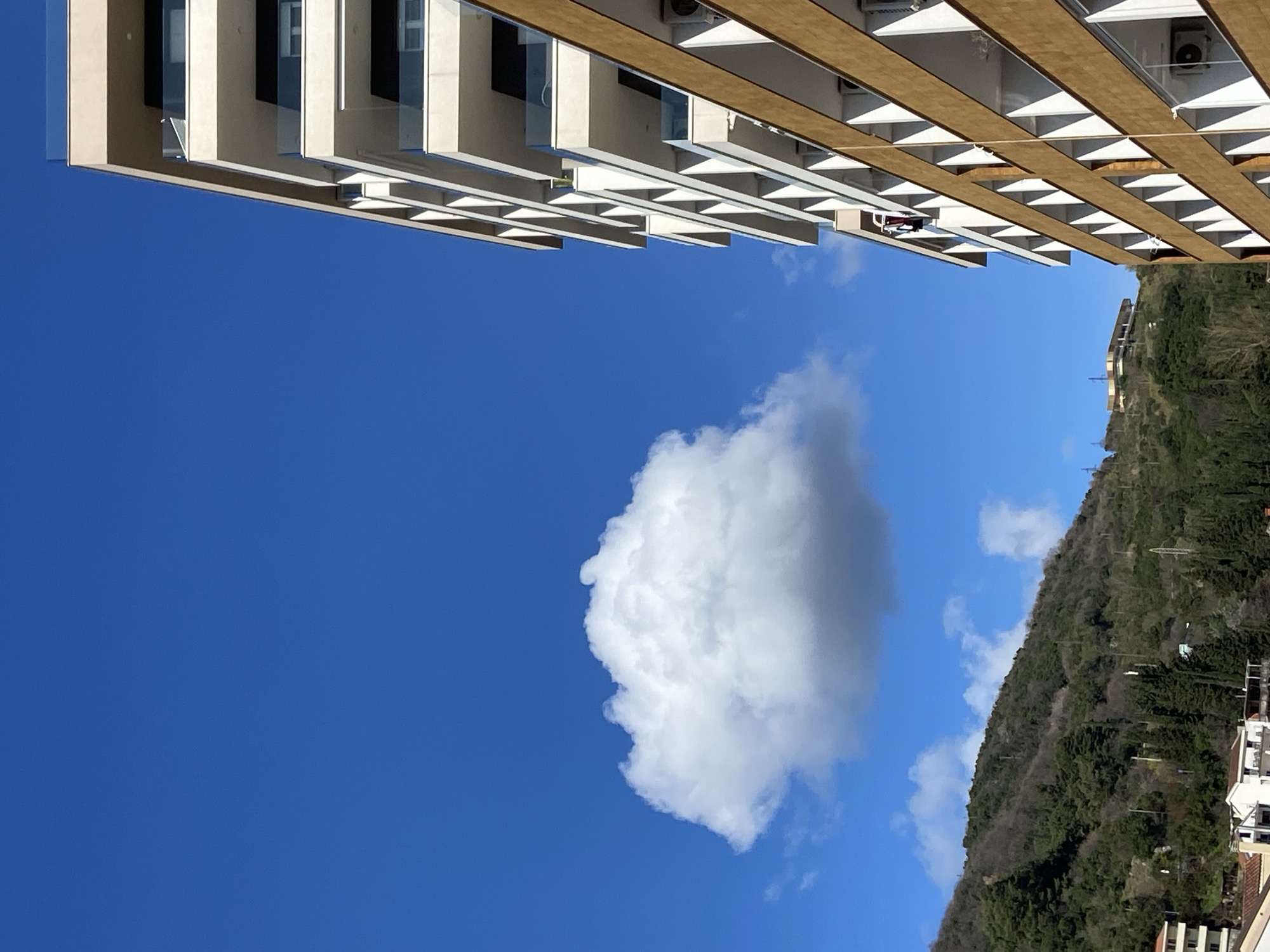 The scene depicts an exterior urban environment in Budva, Montenegro, under a clear, bright blue sky with a prominent, isolated white cumulus cloud positioned in the upper-left quadrant. Two multi-story buildings dominate the right side of the frame. The building in the immediate right foreground presents a light brown facade with numerous recessed balconies, several of which feature external air conditioning units. Adjacent and slightly behind this structure, a taller, modern building with a light beige exterior and tiered balconies equipped with clear glass railings is visible. On an upper-level balcony of this taller, modern building, a partially discernible individual is present, with a distinct red and white textile item, consistent with laundry, draped over the balcony railing. The left and central background are dominated by a large, verdant mountain slope, densely covered in a mix of evergreen and deciduous trees. Several utility poles and a distinct, light-colored building with a flat roof are visible on the upper slopes and distant peak of the mountain.