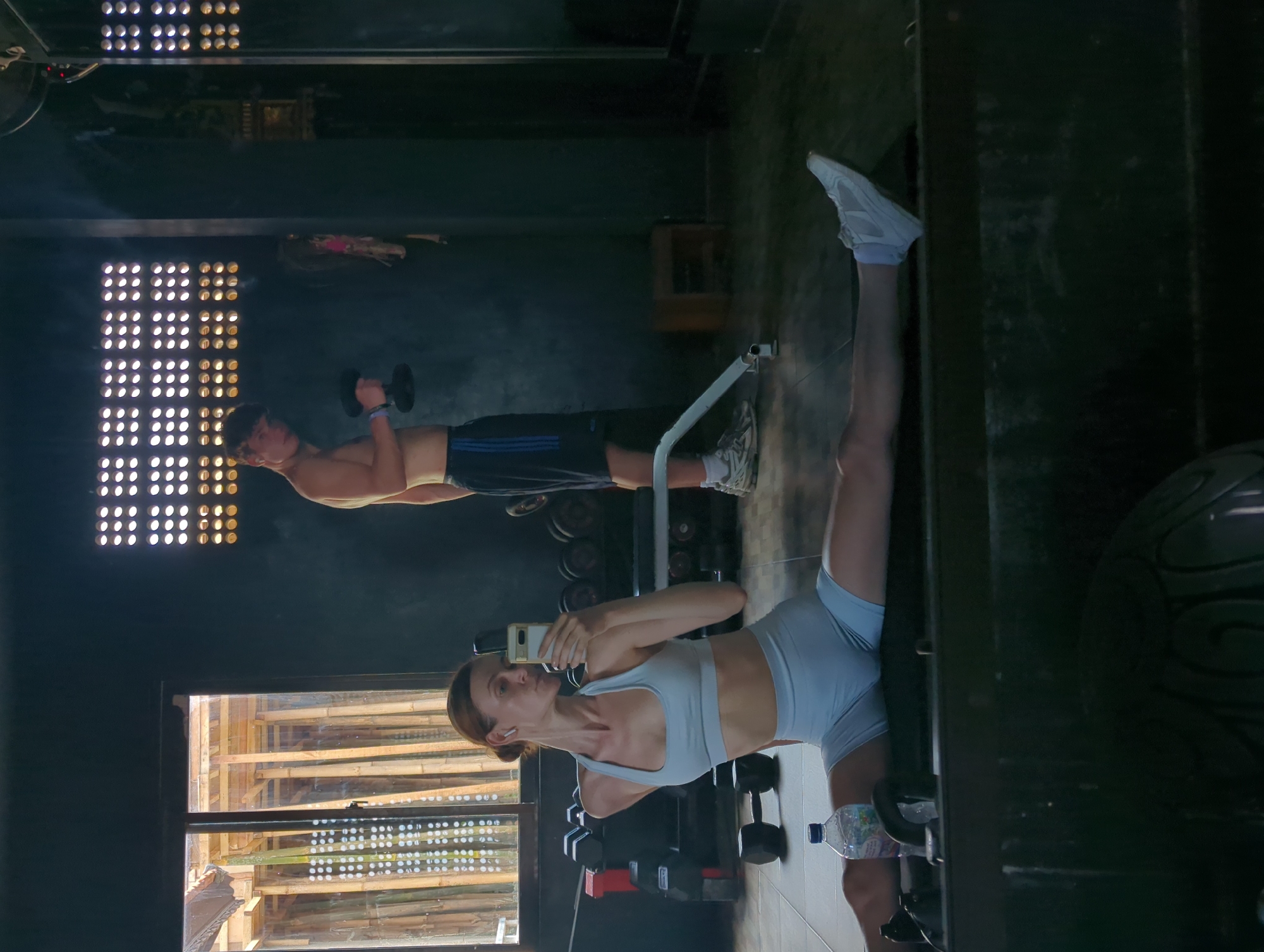 The image shows a mirrored scene inside what appears to be a gym. A woman in light blue workout attire is taking a selfie with her smartphone in front of a mirror. She is standing, with one leg straight up, the other on the ground. Behind her and reflected in the mirror, a man is doing bicep curls with dumbbells. The background includes a wall with rectangular patterns of small circular openings, possibly vents. A water bottle and weights are visible near the woman. The location is Mengwi, Indonesia, suggested by building construction details visible through a window.