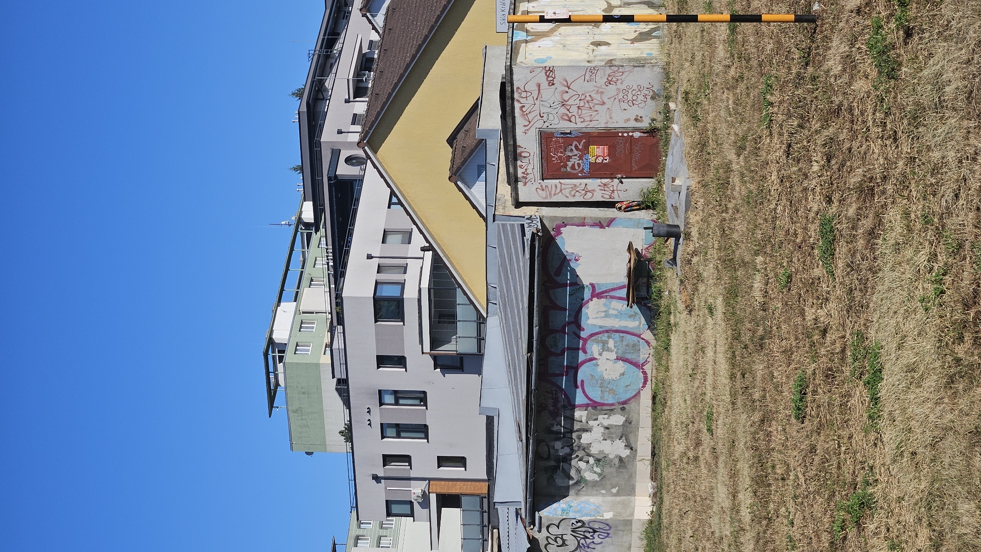 The image depicts an outdoor scene under a clear blue sky, likely in the city of Prešov, Slovakia, given the presence of a street sign partially visible on the right that reads "Sala Kráľova". In the foreground, a dry, overgrown grassy field occupies the bottom half of the frame. In the middle ground, there are several low-rise structures, appearing to be garages or storage units, extensively covered in graffiti. These structures have corrugated metal roofs and concrete walls. One unit has a red metal door. Behind these structures, a complex of modern apartment buildings rises, featuring a mix of light-colored facades, windows, and balconies. The architecture of the apartment buildings suggests an urban setting. The overall scene conveys a contrast between older, graffitied utility buildings and contemporary residential architecture.