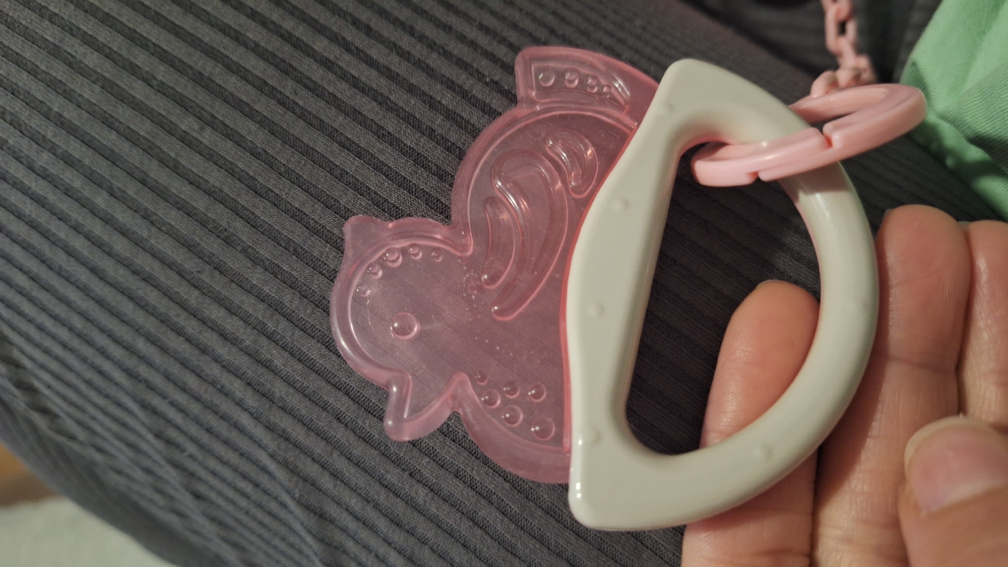 A close-up shot captures a baby teether being held by a person's hand. The main subject is a pink, translucent teether shaped like a stylized aquatic creature, possibly a seahorse or fish. This part of the teether appears to be filled with liquid, as evidenced by numerous small bubbles trapped within the transparent material and intricate raised patterns on its surface.
The pink teether is attached to a white, D-shaped plastic handle, designed for a comfortable grip. This handle features small, raised dots along its edges, providing additional texture. A pink plastic ring or a segment of a chain is linked to the white handle, suggesting it might be part of a larger attachment for securing the teether.
A person's hand is visible in the lower right, with fingers holding the white handle. The background primarily consists of a dark gray, ribbed fabric, likely an article of clothing or a blanket, which has a prominent diagonal stripe pattern. A small portion of light green fabric is also visible in the upper right corner. The lighting indicates an indoor setting, with reflections visible on the smooth plastic surfaces of the teether and handle. No text is visible in the image.