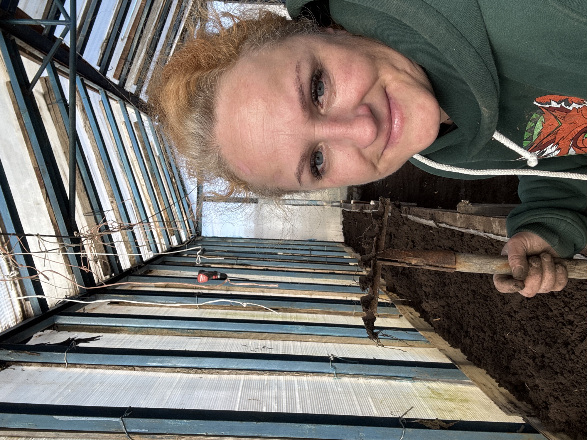 A selfie of a woman in a greenhouse. The woman has fair skin, blue eyes, and reddish-blonde hair pulled back. She is wearing a dark green hooded sweatshirt with a design on the chest. She is smiling. Behind her is the interior of a greenhouse, with a metal and translucent panel roof. She is holding a gardening fork with a wooden handle. The ground is covered in dark soil. A power drill is visible in the background attached to the greenhouse structure. The location is a greenhouse, possibly in a private garden or small agricultural setting. Given the request context, the location is likely in the city of Chaikovsky, Russia.
