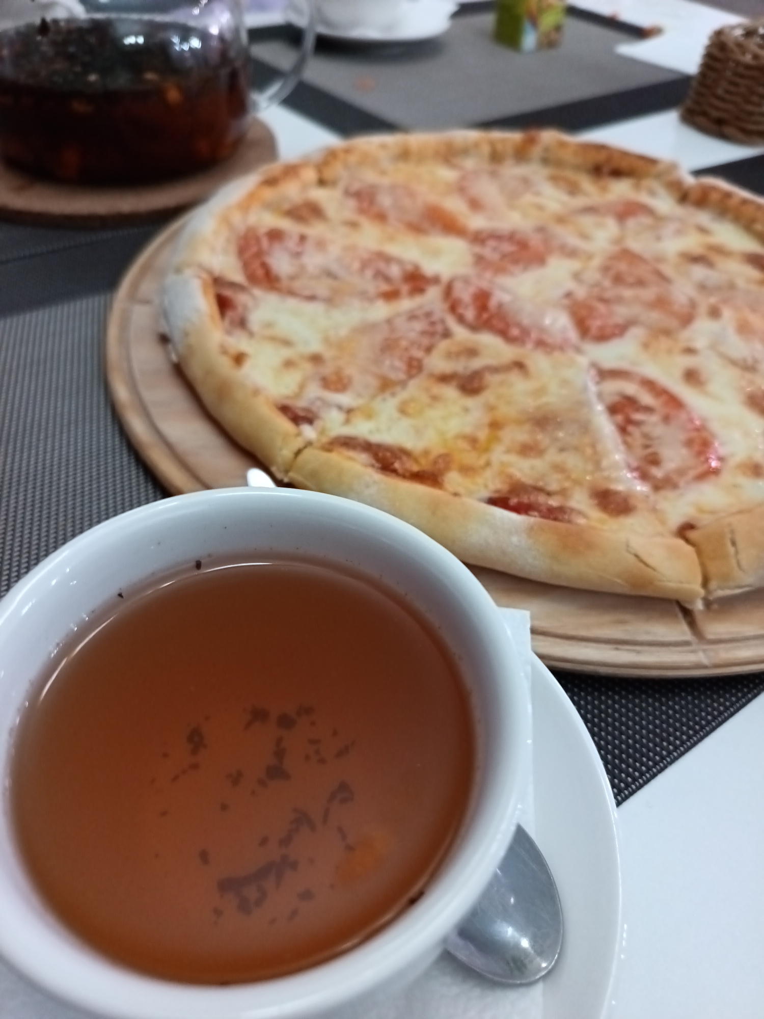 A close-up, high-angle shot captures a meal laid out on a table. In the immediate foreground, a white ceramic teacup filled with dark amber tea sits on a matching white saucer, accompanied by a silver spoon. Small dark specks, consistent with tea leaves, are visible floating on the surface of the tea.
Behind the teacup, a large, round pizza is presented on a wooden serving board. The pizza features a golden-brown, puffy crust and is topped with melted white cheese and slices of red tomato, suggesting a Margherita style. Further in the background, a clear glass teapot or infuser is partially visible, containing a darker liquid with discernible pieces of fruit or tea leaves, indicating freshly brewed tea.
The setting appears to be indoors, possibly a cafe, restaurant, or home dining area in Osa, Russia. The table surface is covered by a placemat with a striped pattern of dark grey and white, next to a plain white surface. Other blurred elements in the background include a small woven basket and a green and white box. The overall scene suggests a quiet moment for a meal or snack. No people or discernible text are visible in the image.