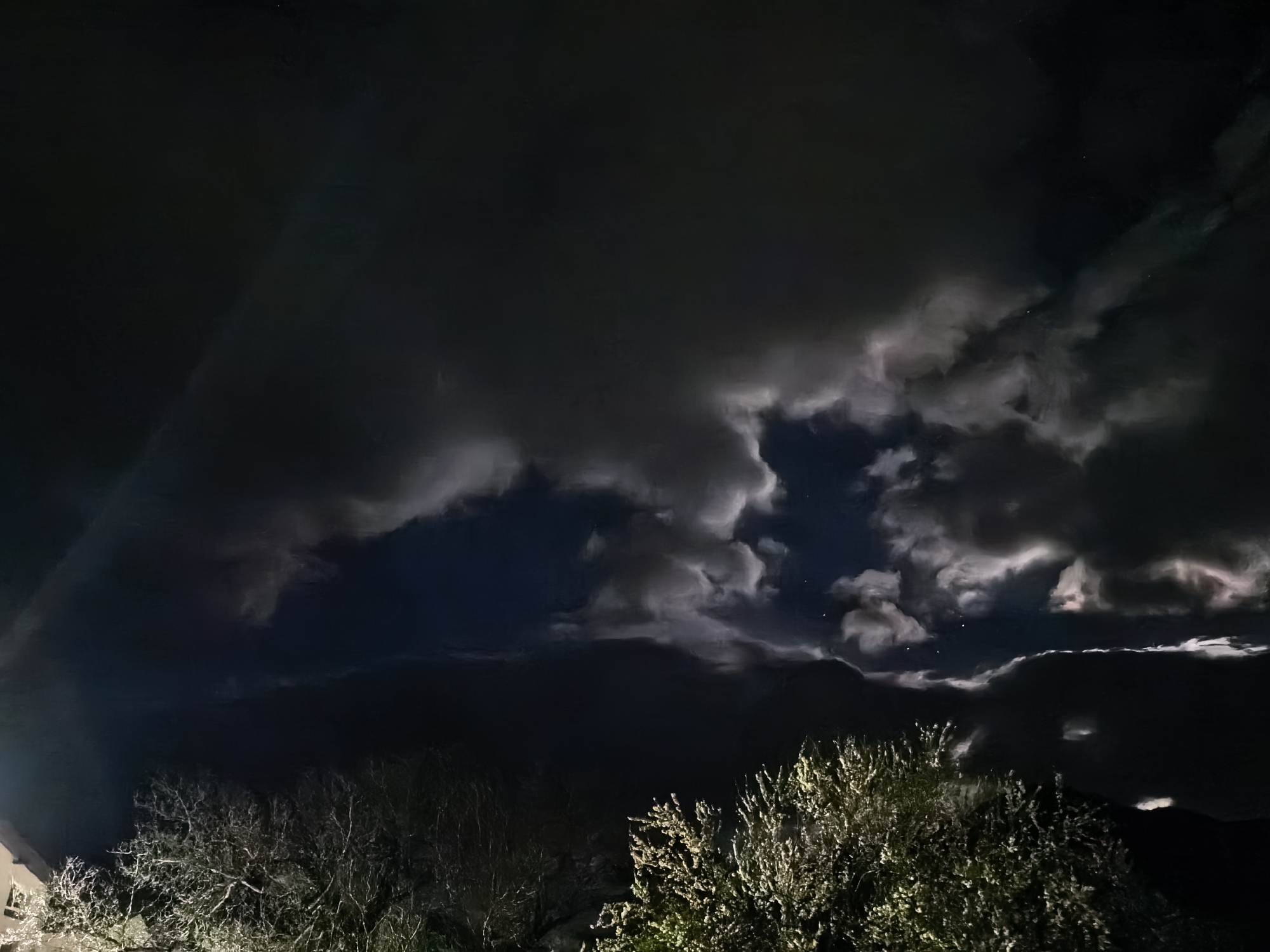 A nighttime scene in Unknown, Unknown depicts dark, heavy clouds dominating the sky. A large opening between the clouds reveals a glimpse of the night sky and a few stars. Below the clouds, silhouetted trees are visible, and light reflects off their leaves. A portion of a building is seen in the lower-left corner. The overall atmosphere is dark and dramatic.