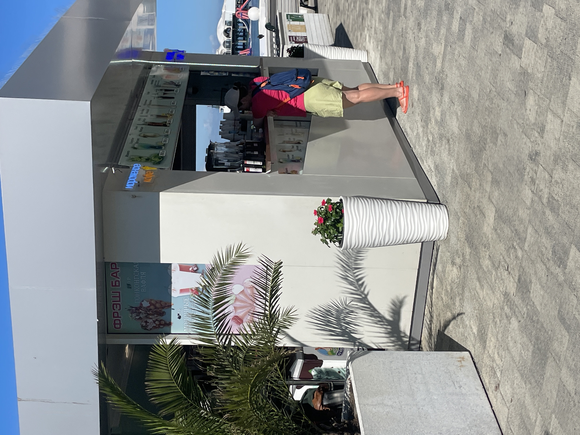 A woman in a pink t-shirt and green shorts stands at a counter in a bright, modern kiosk. She is wearing a white baseball cap and a blue backpack. She appears to be ordering or receiving an item from the kiosk. The kiosk has signs in Russian advertising "FRESH BAR" and "ICE CREAM COFFEE". The kiosk is situated outdoors on a paved walkway. In the background, there are palm trees, potted plants, and a glimpse of buildings and a body of water, possibly the sea, which is consistent with a location in Sochi, Russia. The overall scene suggests a sunny day, likely during a tourist season.