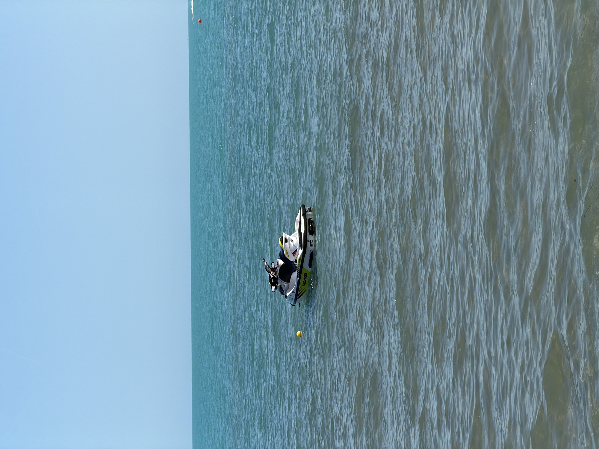 The image depicts a body of water under a clear sky. The sky occupies approximately the top third of the frame, displaying a uniform light blue color with no visible clouds or atmospheric features. A sharp, horizontal line marks the horizon, separating the sky from the water.
The water, filling the lower two-thirds of the image, presents varying shades of blue and green. It appears a deeper turquoise in the mid-ground, transitioning to a slightly murkier blue-green with more visible ripples in the foreground. Small, uniform waves cover the water's surface, indicating calm conditions.
A white, black, and bright yellow personal watercraft (jet ski) is centrally located in the mid-left portion of the water. The brand "Sea-Doo" is visible in yellow lettering on its side. The jet ski is floating stationary, oriented slightly with its stern towards the viewer and its bow pointing towards the right side of the frame. No individuals are present on or near the jet ski.
A small, spherical bright yellow buoy floats on the water to the immediate left of the jet ski. Further in the distance, near the far right edge of the frame and close to the horizon line, a small red spherical buoy is visible. Adjacent to the red buoy, slightly to its left, a faint white line on the water indicates a distant wake or spray, suggesting the presence of another watercraft or recent movement, though the source craft is not discernible. No landmass or other significant structures are visible. The lighting suggests a clear, bright day.