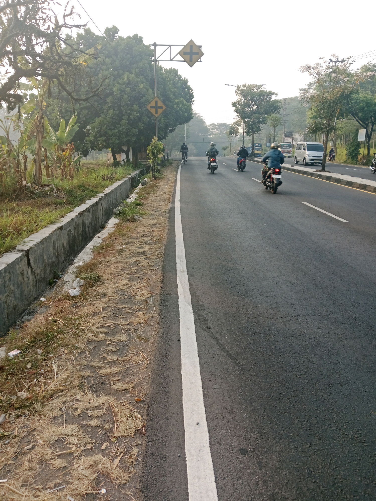 A road scene in Salatiga, Indonesia, shows a two-lane asphalt road with a white line dividing the lanes. On the left side of the road is a concrete drainage ditch with dry grass and some debris. Beyond the ditch, there are banana trees and other vegetation. On the right side of the road, there is a sidewalk with trees and a white van. Several motorcycles are on the road, some carrying riders. Traffic signs are visible, including a yellow diamond-shaped sign with a cross symbol. The sky is hazy, suggesting it may be early morning or late afternoon.
