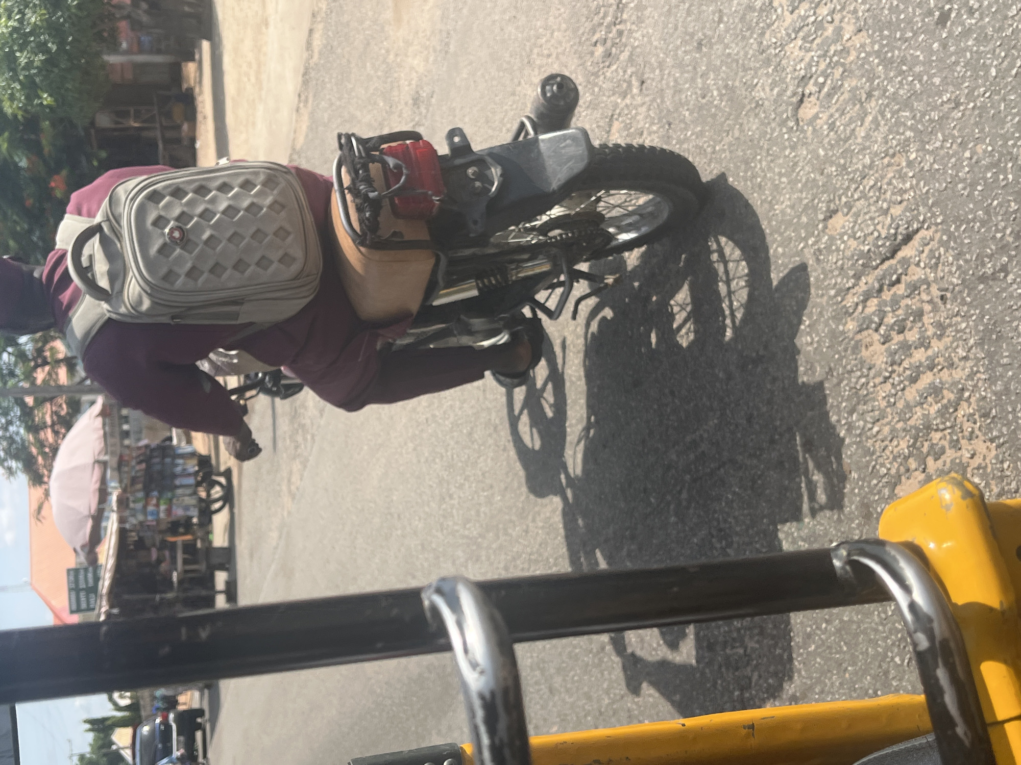 The image captures a scene from Jalingo, Nigeria, looking from the perspective of a tricycle. A person wearing a maroon outfit and a light-colored backpack is riding a motorcycle on a paved road. The motorcycle has a brown seat and a red taillight. A stall with goods displayed under a light pink umbrella is visible on the right side of the road, with trees and buildings in the background. The road surface is textured with small stones and cracks, and shadows of the motorcycle and tricycle are cast on the ground. The lighting suggests it is daytime.