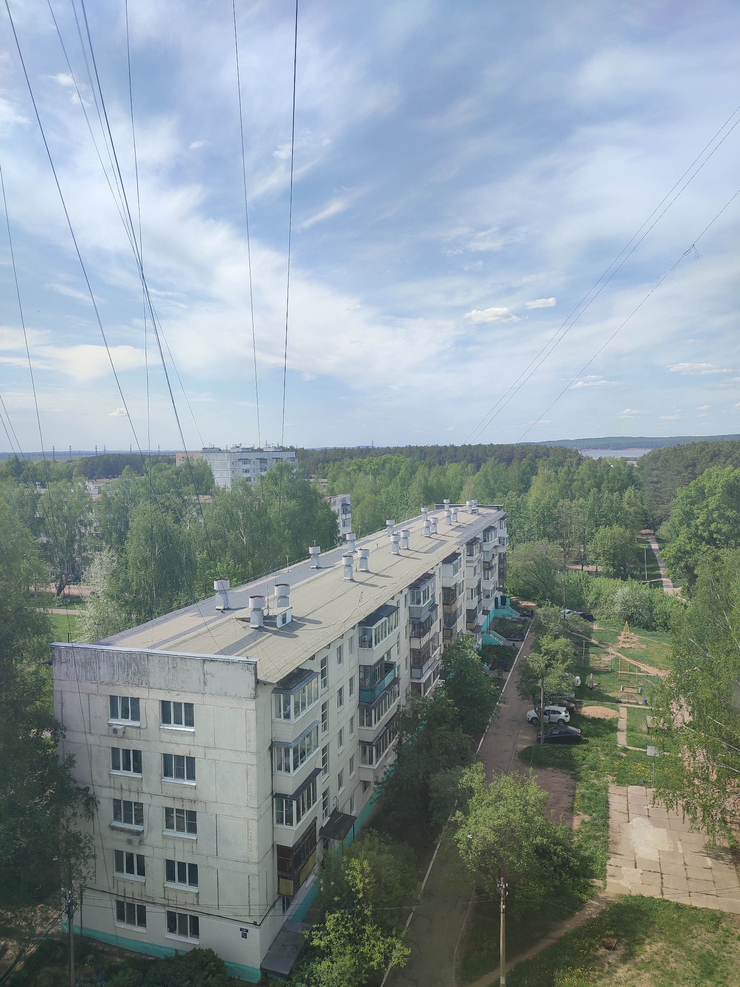 A high-angle shot depicts a multi-story apartment building in the city of Unknown, Unknown, surrounded by trees and greenery. The building has a flat, gray roof with several ventilation shafts or chimneys. Its facade is light gray, with rows of windows. Balconies are visible along the side of the building.
Electrical or telephone wires crisscross the sky above the building.
A road runs along the side of the building, lined with parked cars and trees. A playground is also visible. In the distance, more buildings and trees can be seen, with a body of water visible between the trees.
The sky is partly cloudy, with patches of blue visible. The overall light suggests it is daytime.
