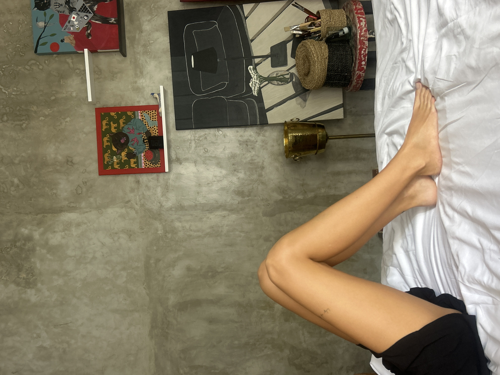 A woman's legs and feet are seen lying on a white bed in a room with a concrete wall. The lower part of the woman's body is visible, showing she is wearing black shorts. The room is decorated with artwork, including a picture of a cat, a portrait of a chair and side table, and another picture. A vintage lamp can be seen in the corner near the bed. The location is likely North Kuta, Indonesia.