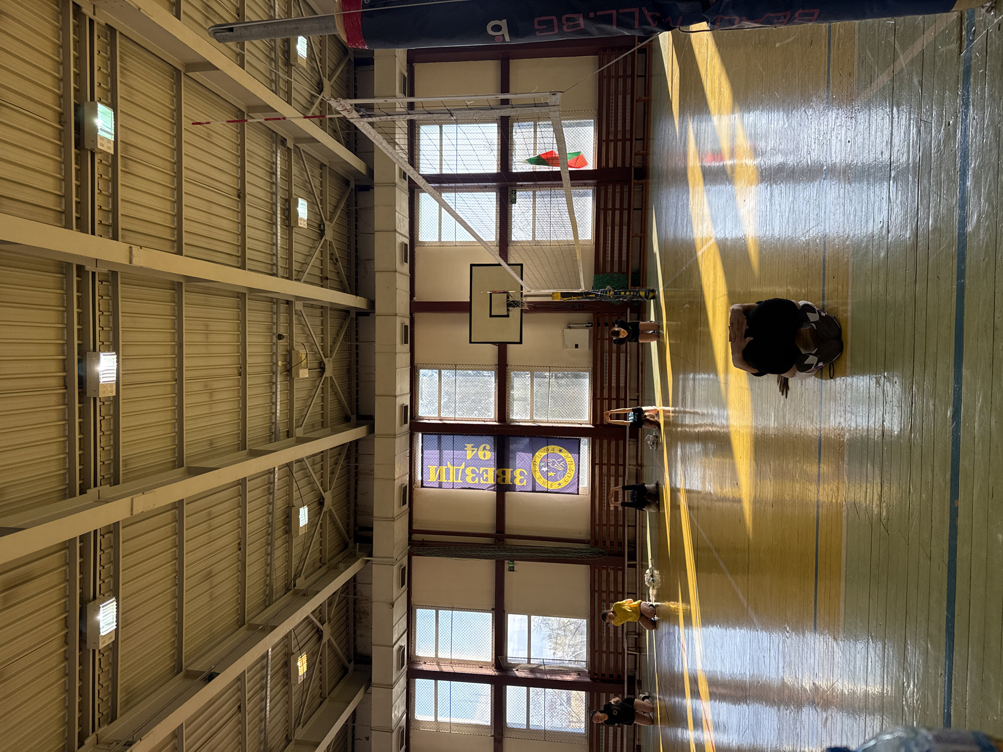 The image is an interior shot of a gymnasium, possibly located in Sofia, Bulgaria, as indicated by the Cyrillic text "ЗВЕЗДИ 94" on a banner, translating to "Stars 94." The gym appears to be well-lit with natural light coming through a series of windows along one wall. These windows are rectangular and separated by dark brown dividers. Below the windows is a basketball hoop. A volleyball net is suspended from the ceiling. The floor is wooden with yellow and blue painted lines marking out the sports court. Several people are visible on the court, engaged in some kind of activity. They appear to be wearing athletic attire. The roof of the gym consists of metal panels, supported by a grid of structural beams and equipped with multiple light fixtures.