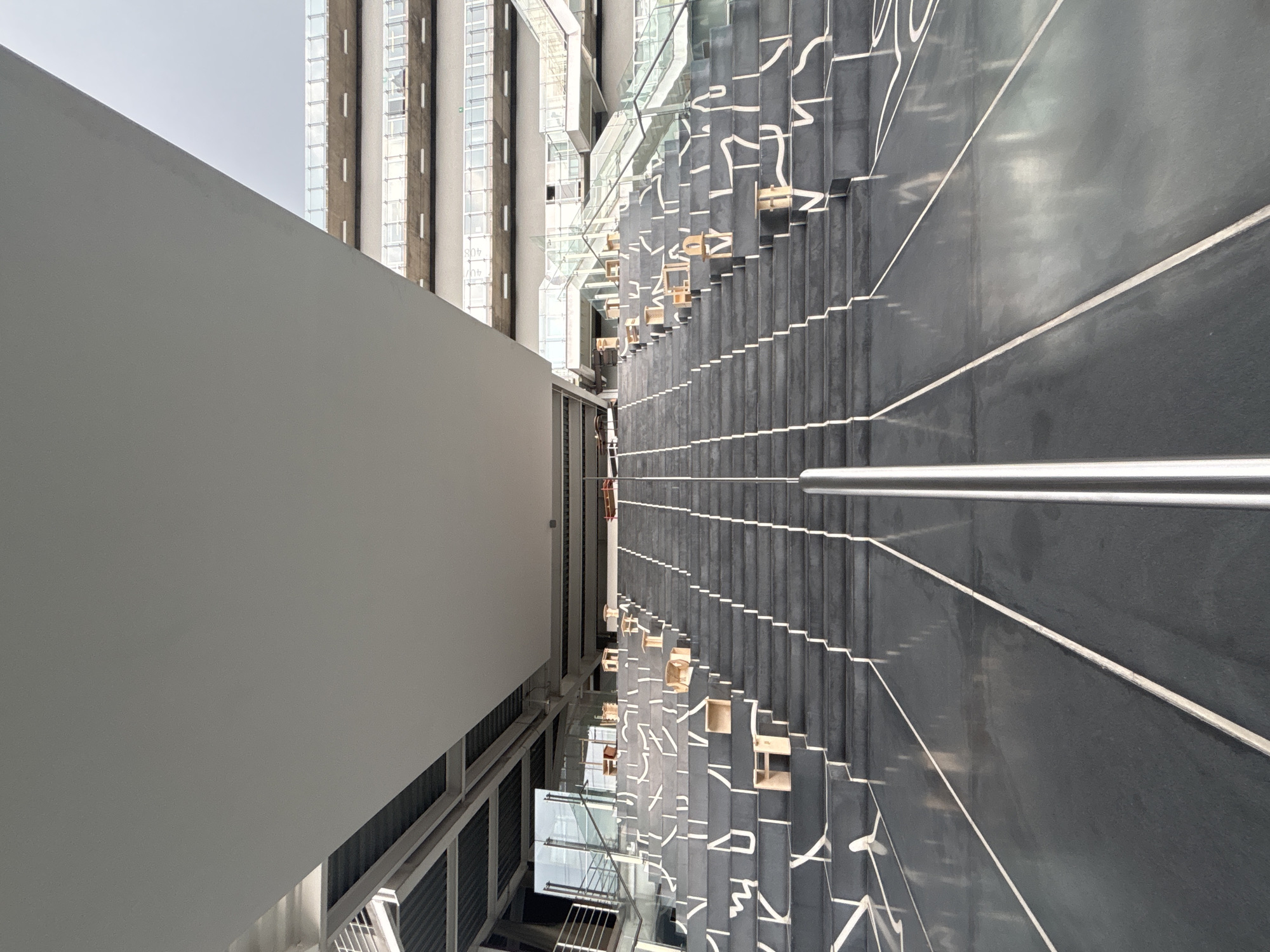 This is a low-angle, full shot of a grand, outdoor staircase leading upwards towards a modern building. The staircase is constructed from dark gray stone and features white geometric lines painted across the risers, creating a visually striking pattern. Small, light-colored wooden cuboids are placed sporadically on some of the steps, serving as decorative elements or perhaps informal seating.
A central, polished metal handrail runs up the middle of the stairs, dividing them into two symmetrical ascending paths. The steps are wide and numerous, disappearing into the distance. Above the staircase, a large, flat, light-colored overhang or ceiling casts a shadow, partially obscuring the upper levels of the building.
In the background, the modern building is visible, characterized by multiple stories with balconies and glass railings. The overall aesthetic is contemporary and architectural. The sky above is overcast and appears muted, suggesting a cloudy day. No people are visible in the immediate foreground or on the staircase.
The location is the UNAM Central Library, also known as the Biblioteca Central, in Mexico City, Mexico. This iconic structure is renowned for its striking mural by Juan O'Gorman, which is integrated into the building's exterior. While the mural itself is not directly visible in this shot, the architectural style and grand scale are consistent with the UNAM campus.