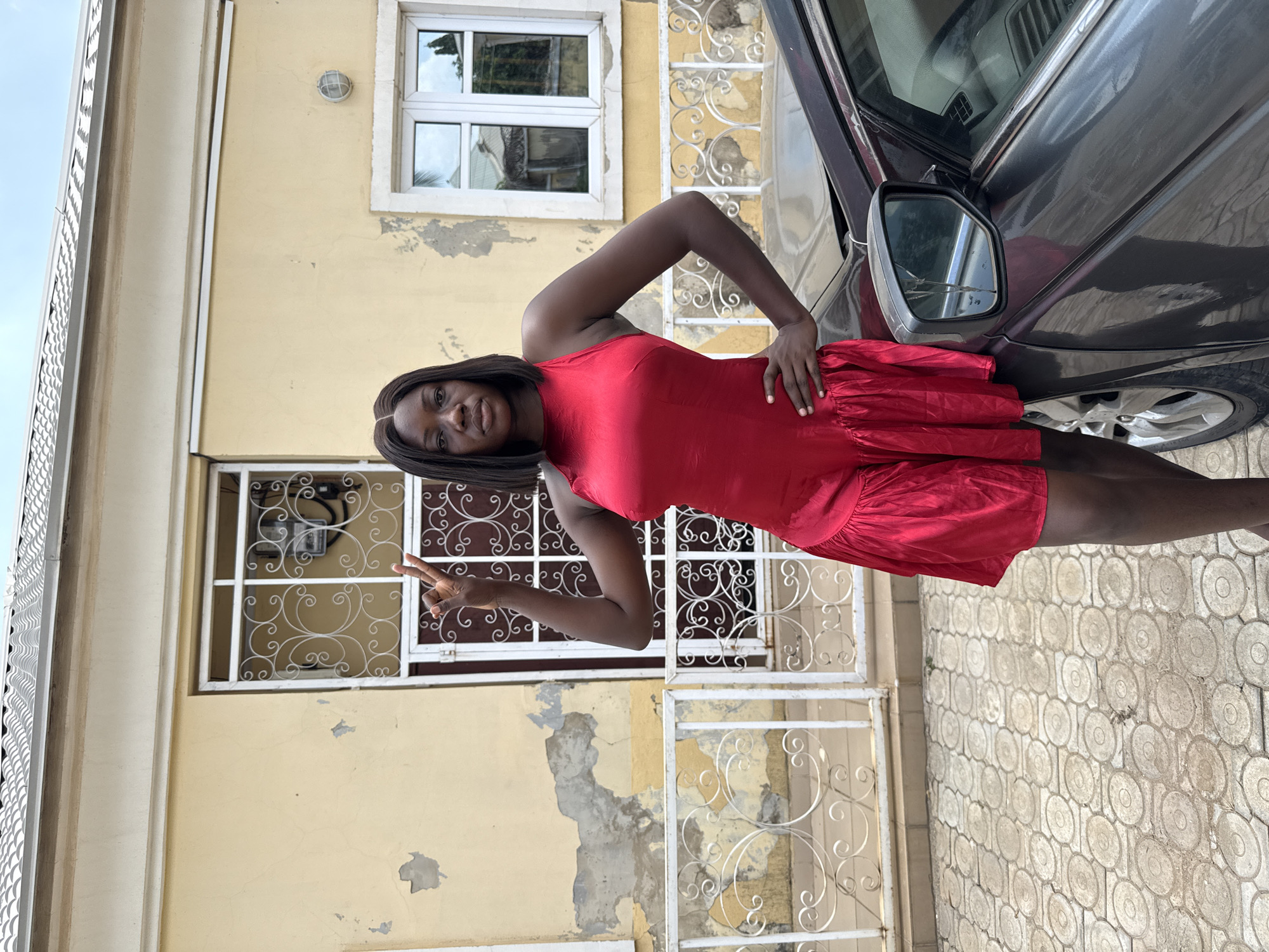 A medium shot shows a Black woman in a red dress standing outside in front of a building and next to a car. The woman is positioned on the right side of the frame. She has black hair and is wearing a red dress. She is making a peace sign with her left hand and has her right hand on her hip.
The building is yellow and has white window frames with decorative metal bars. The paint is peeling in several places. A car, mostly gray, is on the right side of the frame, partially blocking the view. The car's side mirror is visible. The ground is covered in light gray patterned paving stones. Based on the architecture and environment, the location is likely in a residential area, potentially in Abuja, Nigeria.