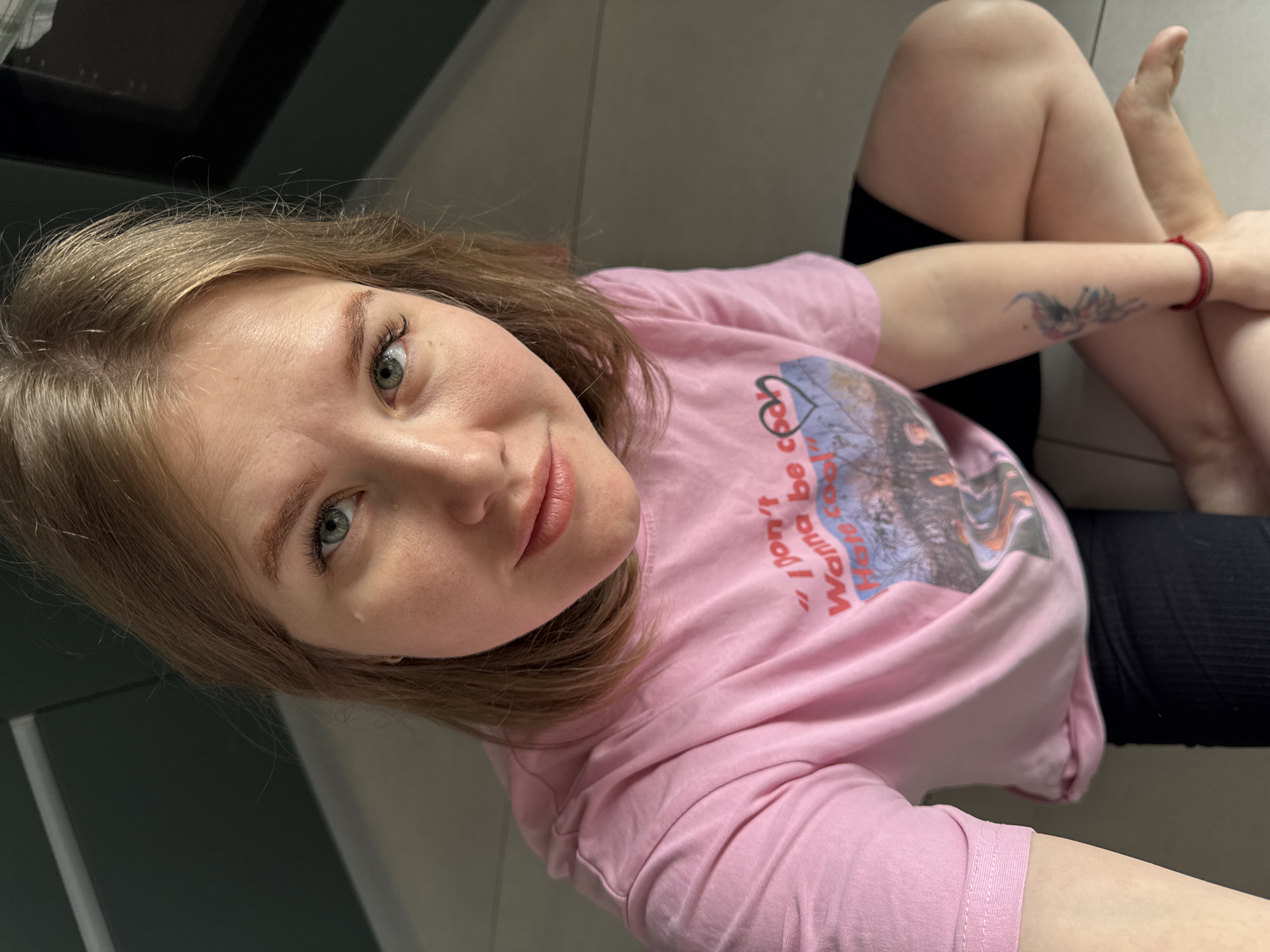 A female individual is depicted sitting on a light-tiled floor indoors in Gornyi (Dvurechenskoe S/P), Russia. She has light brown hair, green eyes, and fair skin, looking directly into the camera. She wears a pink short-sleeved t-shirt with a graphic design featuring text that partially reads "I don't Wanna be cool" and an illustrated landscape. Her lower body is clad in black shorts or leggings, with her legs folded beneath her. A multi-colored tattoo, appearing to be a mythical creature, is visible on her left forearm, and a red string bracelet is on her left wrist. The background includes a dark-colored wall with a horizontal light line and a partially visible dark, reflective rectangular object.