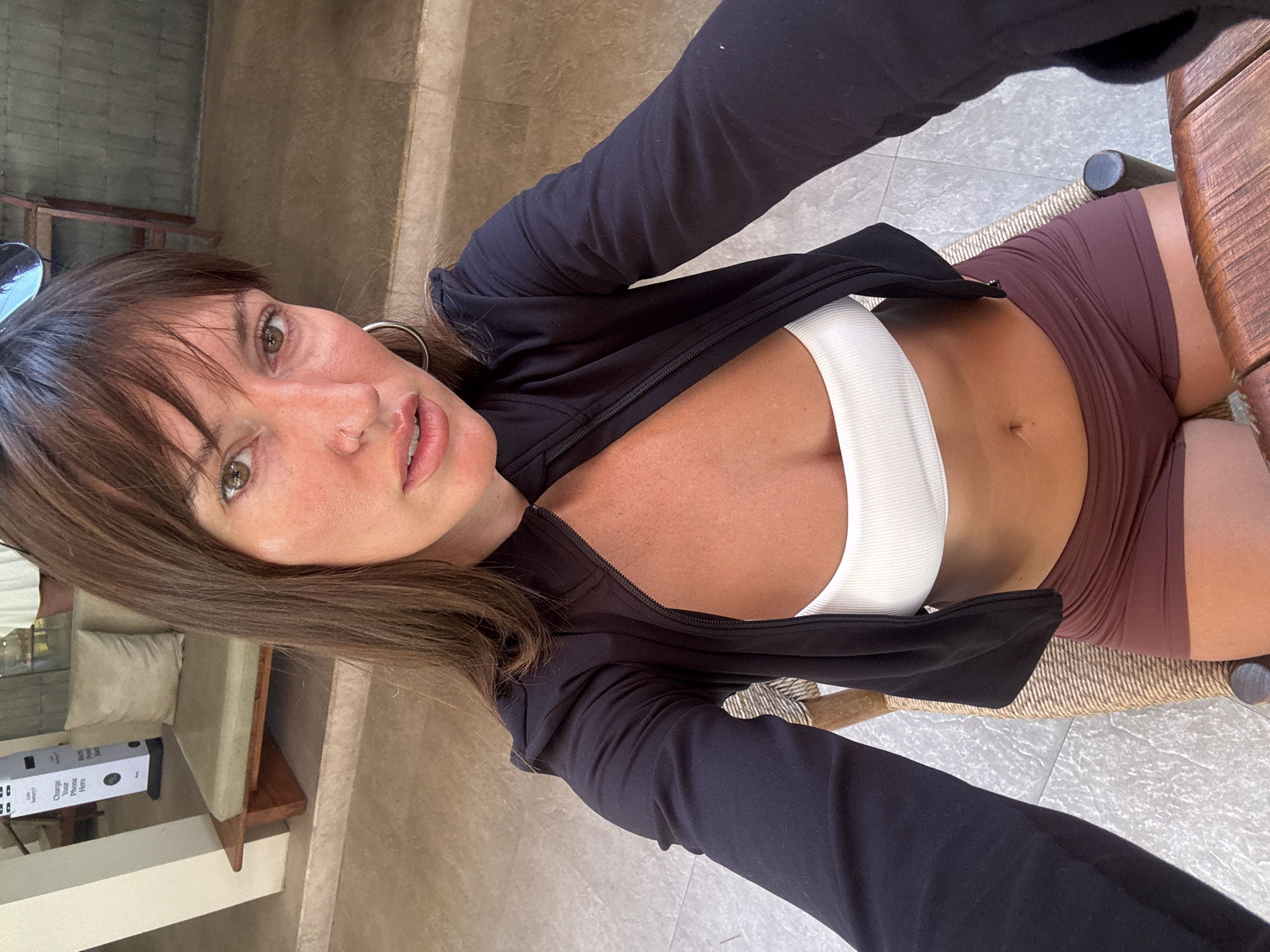 A young Caucasian woman with brown hair and bangs is taking a selfie in a seated position. She is wearing a white bandeau top and dark reddish-brown shorts, with an unzipped black jacket over her shoulders. Her left arm is extended towards the camera. The background suggests an outdoor or semi-outdoor setting, possibly a cafe or restaurant, with a stone tiled floor, a wooden table in the foreground, and a woven chair. In the background, there's a bench with a light green cushion and a white stand with text, possibly a charging station. The lighting is natural and the image appears to be taken during the day.