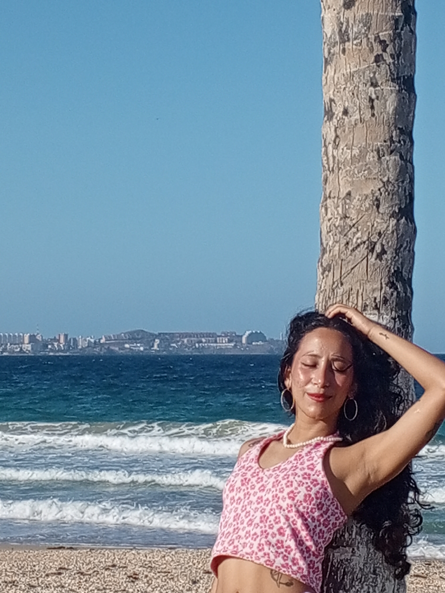 A woman is posing on a beach next to a palm tree. She is wearing a pink and white floral top, hoop earrings, and a pearl necklace. She is leaning against the tree with her eyes closed and one hand on her head. The beach has white pebbles. The ocean has small waves breaking on the shore. In the distance, the skyline of a city is visible, which is likely Porlamar, Venezuela. The sky is clear and blue.