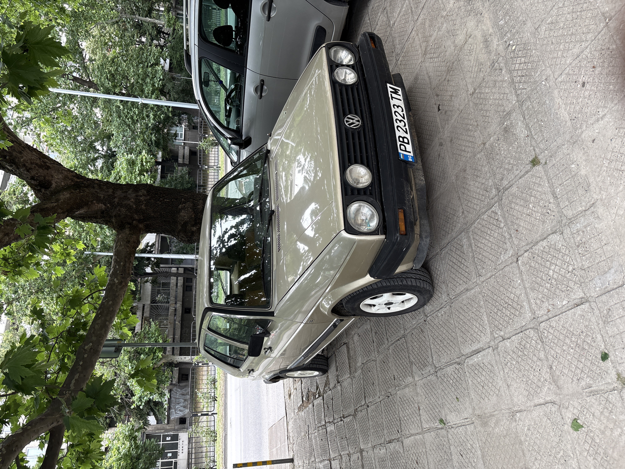 A parked, beige Volkswagen Golf Mk2 is observed on a sidewalk in Plovdiv, Bulgaria, with a silver hatchback parked to its right. The Golf faces slightly left, angled towards a street. Its front license plate reads "PB 2323 TM". The sidewalk is paved with square concrete tiles, some of which are cracked. A mature tree with large leaves is positioned behind the Golf, casting dappled shade on the car and sidewalk. In the background, a concrete building with numerous windows and some graffiti is visible across the street, along with a black metal fence. The environment appears to be an urban setting, likely a residential street, given the presence of trees and buildings. The scene is static, with no people or active events depicted.