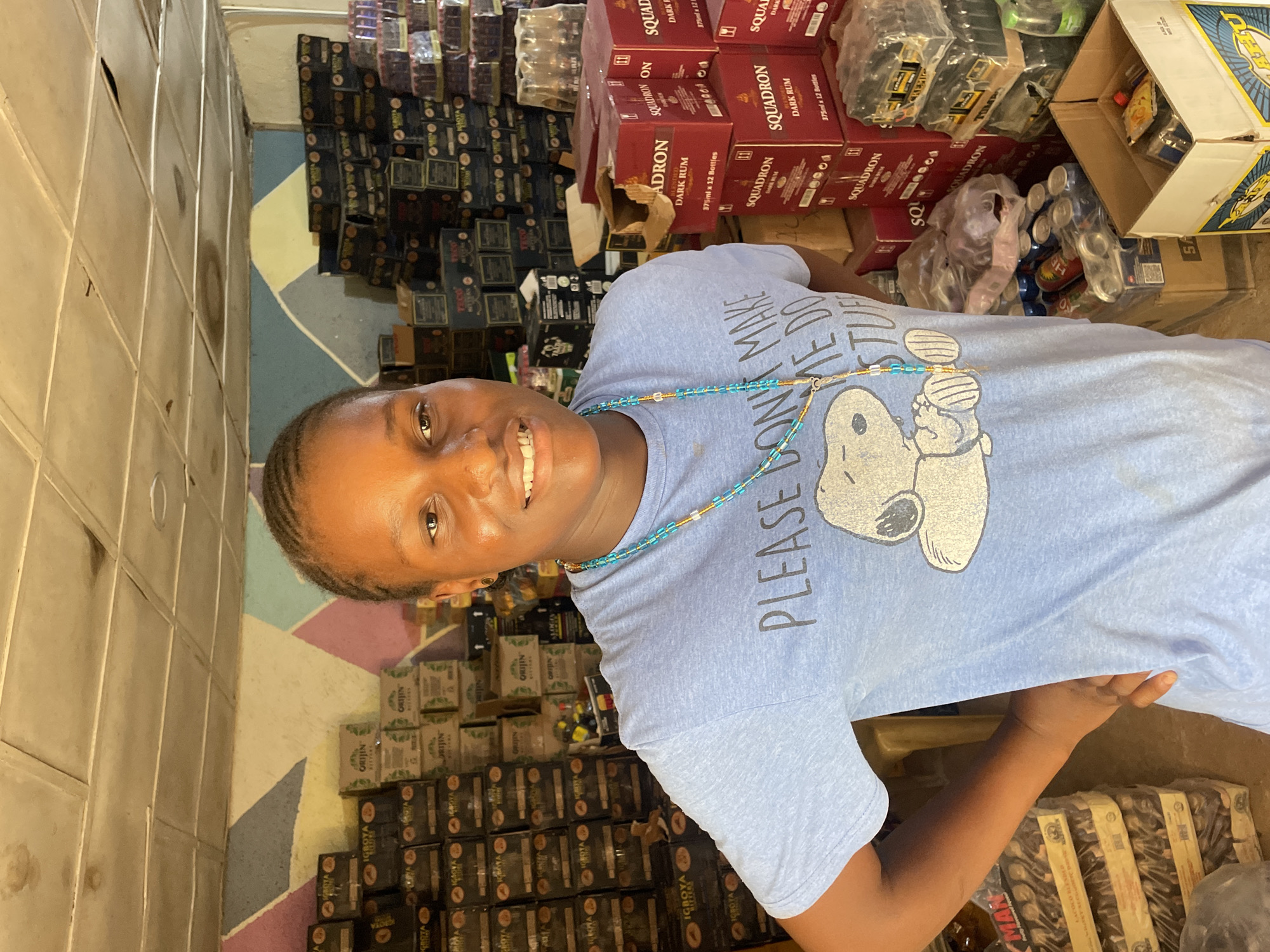 The image features a smiling young Black person, likely female, captured in an indoor setting that appears to be a shop or storeroom in Jos, Nigeria. The individual is positioned in the foreground, looking directly at the camera with a wide, friendly smile. They have short, braided hair and are wearing a light blue t-shirt. The t-shirt bears the text "PLEASE DON'T MAKE ME DO STUFF" alongside a cartoon illustration of Snoopy. A multi-colored beaded necklace, predominantly in shades of turquoise, blue, and brown, adorns their neck.
The background is densely packed with stacked boxes and packaged goods, creating the impression of a retail or storage space. Prominent items include numerous red boxes clearly labeled "SQUADRON DARK RUM," with additional information stating "375ml x 12 Bottles." Other visible boxes carry brand names such as "ORIJIN" and "IGBOYA BITTERS." Partially visible text on other boxes includes "CHIEFTAIN," "VAG," "MAN," and "ACOCO BLENDLO." Some goods are wrapped in clear plastic. The floor beneath the person and the stacked goods features a distinctive geometric pattern in light blue, white, and purplish-brown hues. The wall to the left is textured, possibly tiled, in a neutral color. The scene depicts a common commercial environment in a local context.