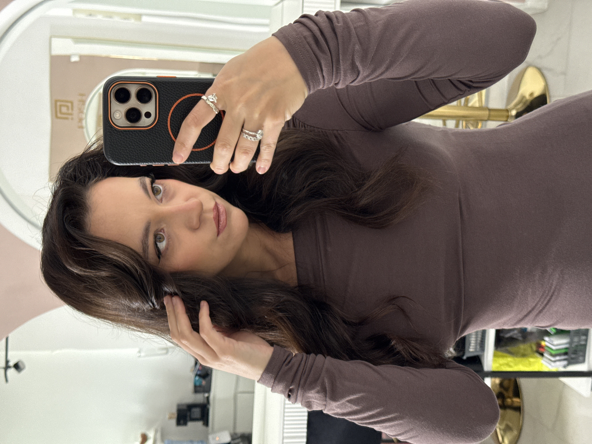 The image shows a woman taking a mirror selfie in what appears to be a salon or boutique. She has long, dark brown hair, is wearing a mauve long-sleeved top, and is wearing rings on her fingers. Her right hand is holding a black phone with an orange trim, which is reflecting in the mirror. In the background, the mirror reflects a sign with a logo that says "POSH." The setting appears to be indoors with white walls and some shelving units visible. There are no elements in the image that indicate the location is in Kazan, Russia.