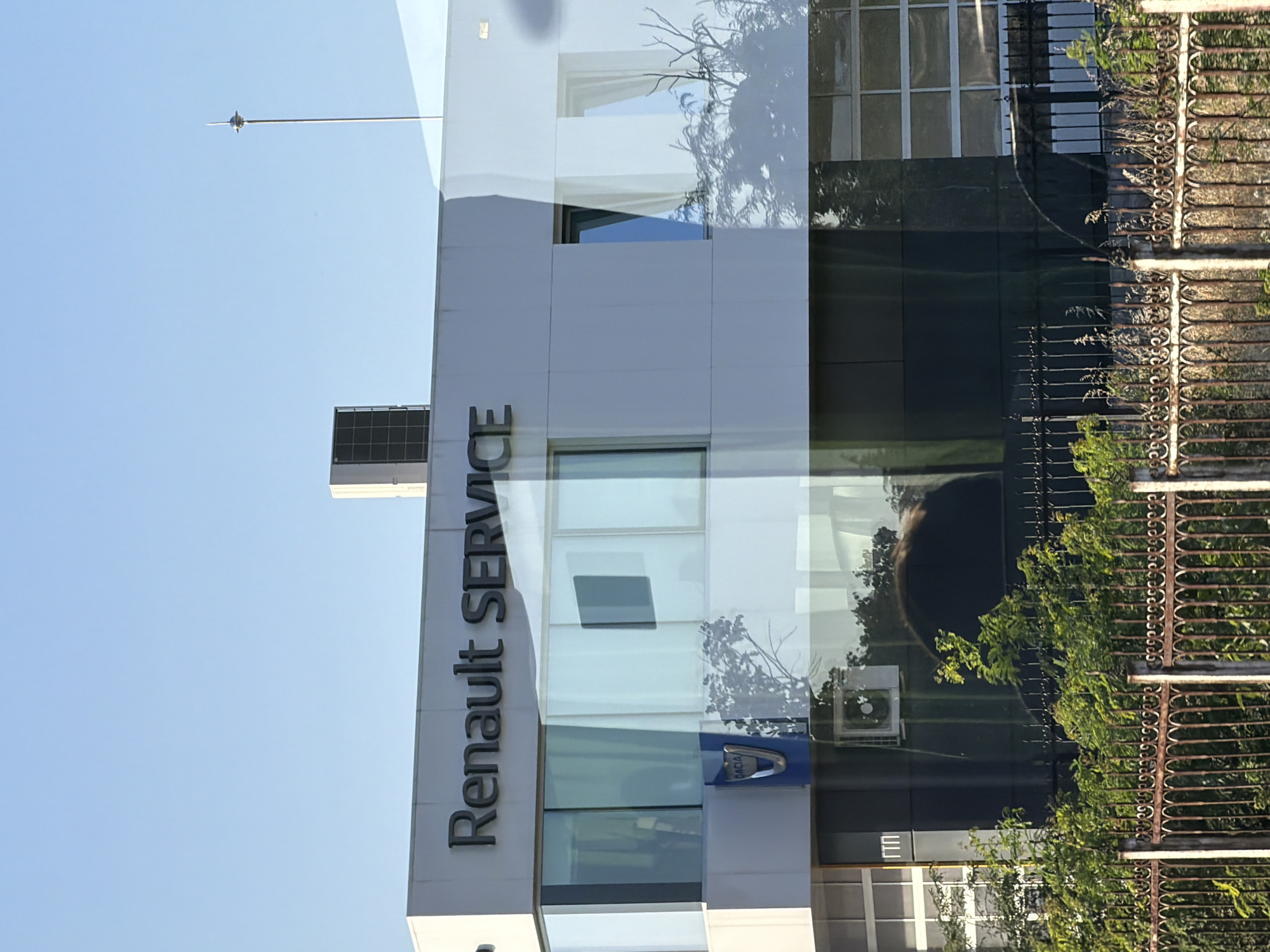 The image shows the exterior of a building with "RENAULT SERVICE" signage prominently displayed in dark lettering against a white facade. Below the sign, there are large glass windows reflecting the sky and surrounding trees. A Dacia logo is visible on a blue background to the left, below the windows. The building appears to be a commercial establishment, likely a car dealership or service center. A rusty metal fence with intricate scrollwork and green foliage in front of it partially obscures the lower part of the building. The sky is clear and blue, indicating daylight. This scene is located in Plovdiv, Bulgaria, a well-known city with a rich history and a modern commercial landscape, which includes numerous automotive service centers like this one.
