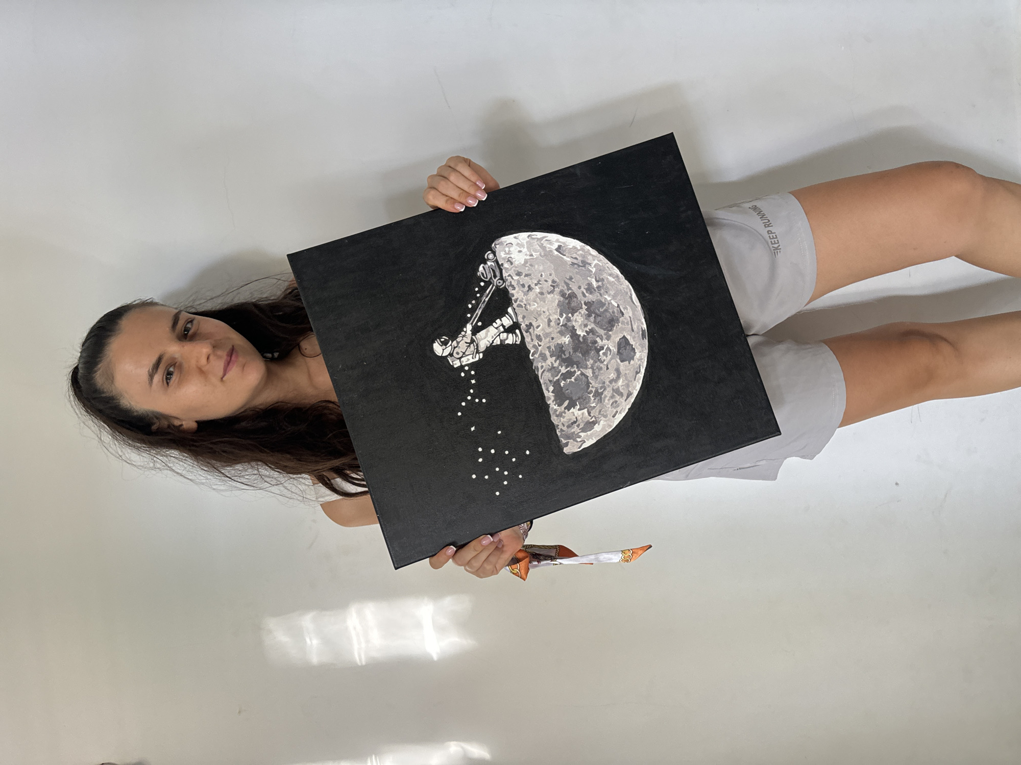 A woman is holding a square painting in front of her. The painting depicts a crescent moon against a black background, with a figure in a spacesuit appearing to be mining the moon. Below the figure are small white dots. The woman has dark hair pulled back, and she is wearing light gray shorts with "KEEP RUNNING" printed on the left leg. In her right hand, she is holding a patterned object. The background is a plain white wall. The photograph appears to have been taken indoors. Location information (Mũi Né, Vietnam) is not evident from the image itself.