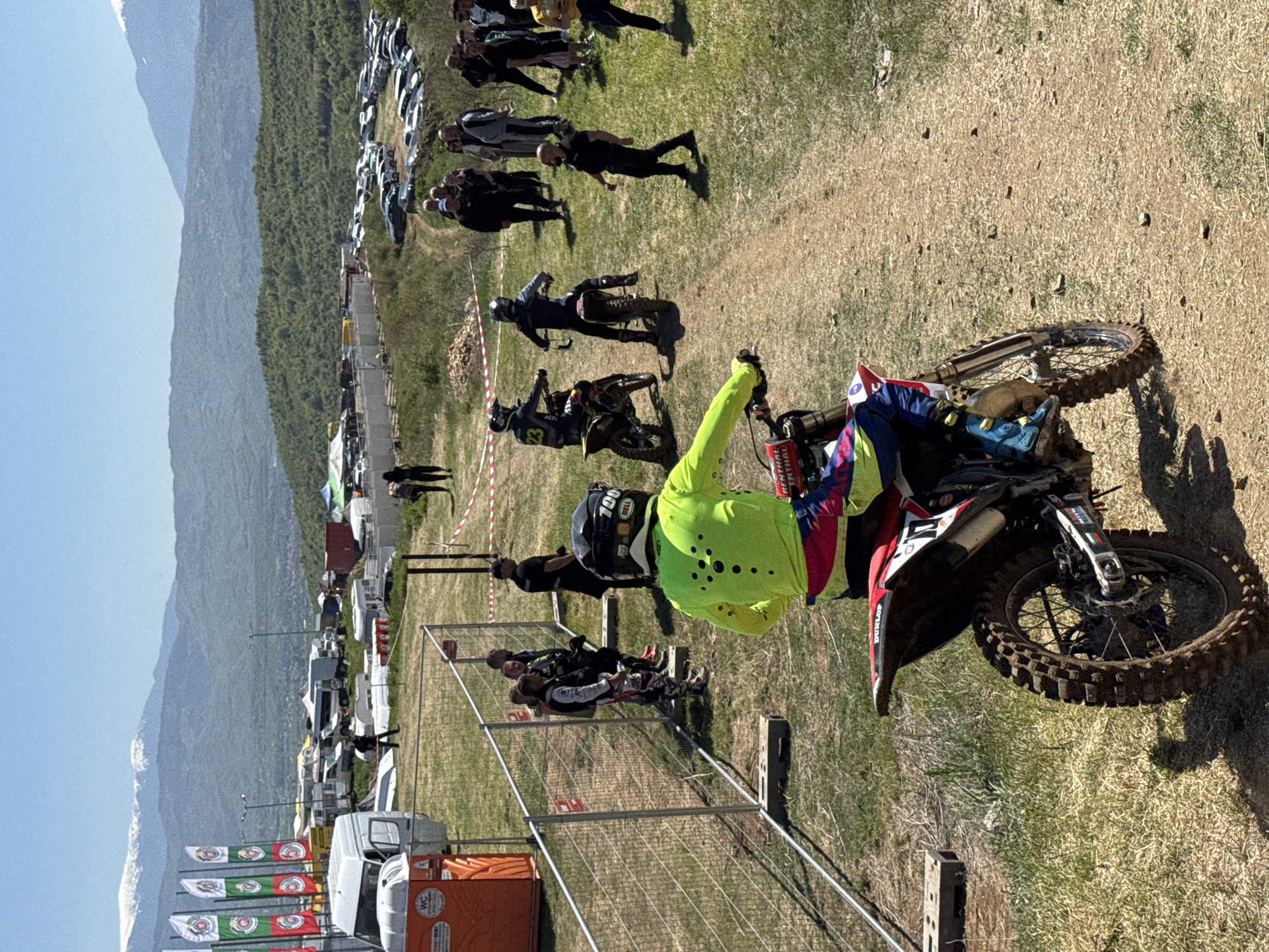 The image shows a motocross event in Selishte, Bulgaria. A motocross rider in a yellow jacket is on a dirt bike at the starting line, facing right. Other riders are lined up behind him. Spectators are visible on the left, separated from the track by a wire fence and red and white striped tape. Mountains are visible in the background. Vehicles, tents, and flags are also present.