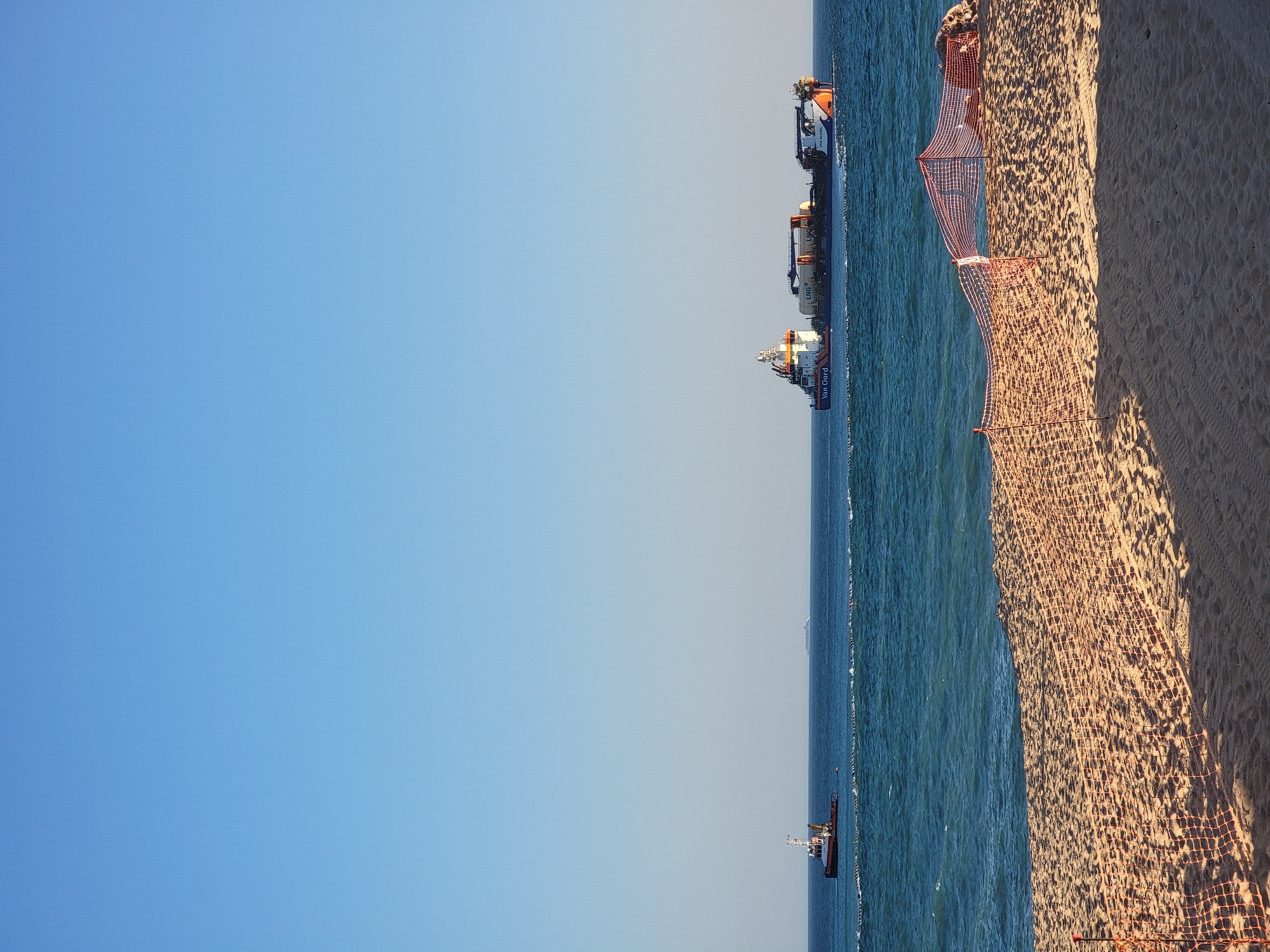 An orange safety net is erected on a sandy beach in Quarteira, Portugal. The net is the foreground of the image. Beyond the beach, the blue sea is calm with gentle waves. In the middle ground, two vessels are present on the water. On the left, a smaller tugboat is moving away from the camera, and on the right, a larger, multi-part vessel is sailing towards the right, displaying the text "Van Oord" and "VOX ALEXIA" on its side. Both vessels are sailing under a clear blue sky.